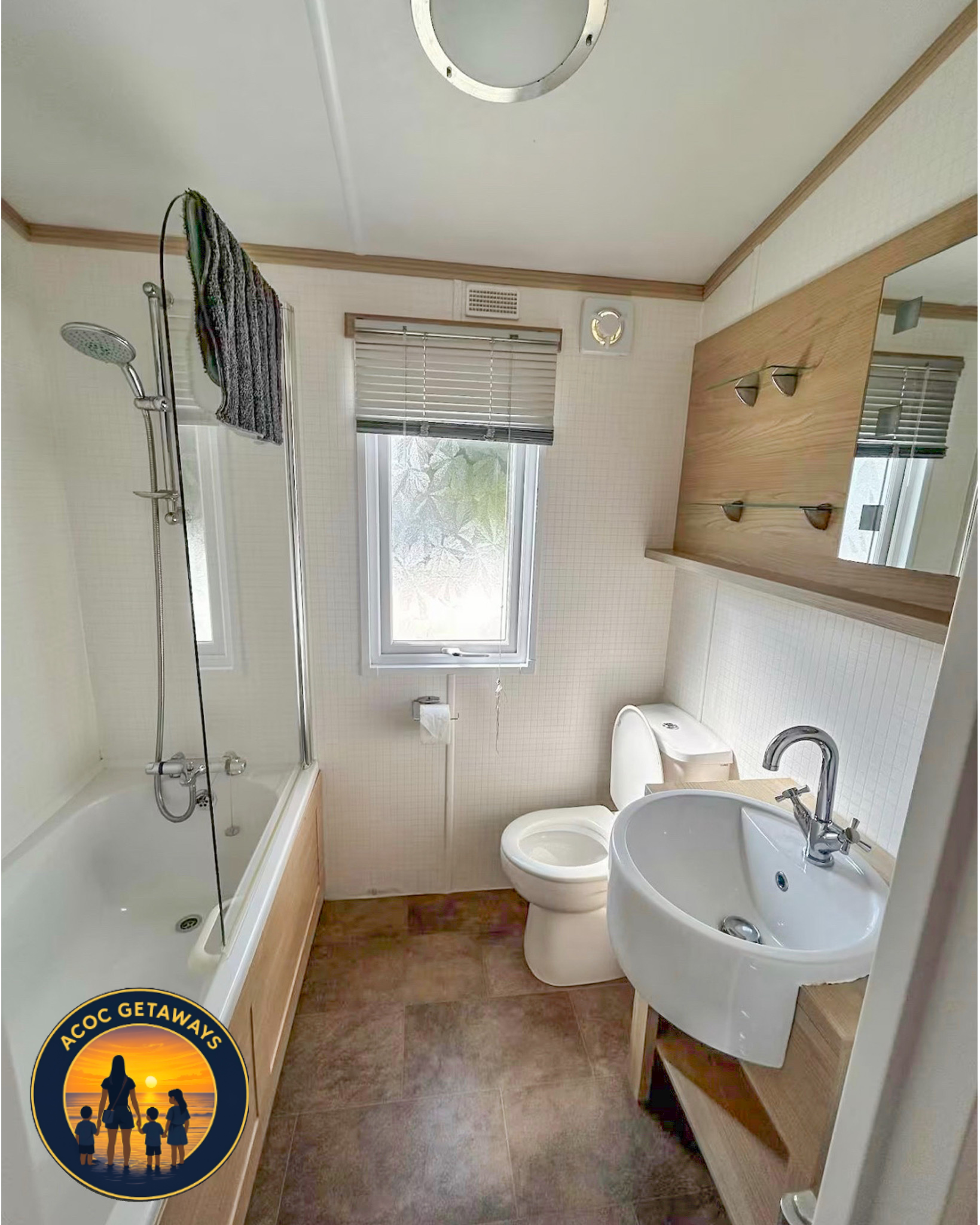 A small bathroom with a shower, toilet, and sink. The shower has a glass door and towels hanging on a rail. There is a window with blinds and a small circular vent. The floor is brown, and the walls are light with a wooden shelf and mirror above the 