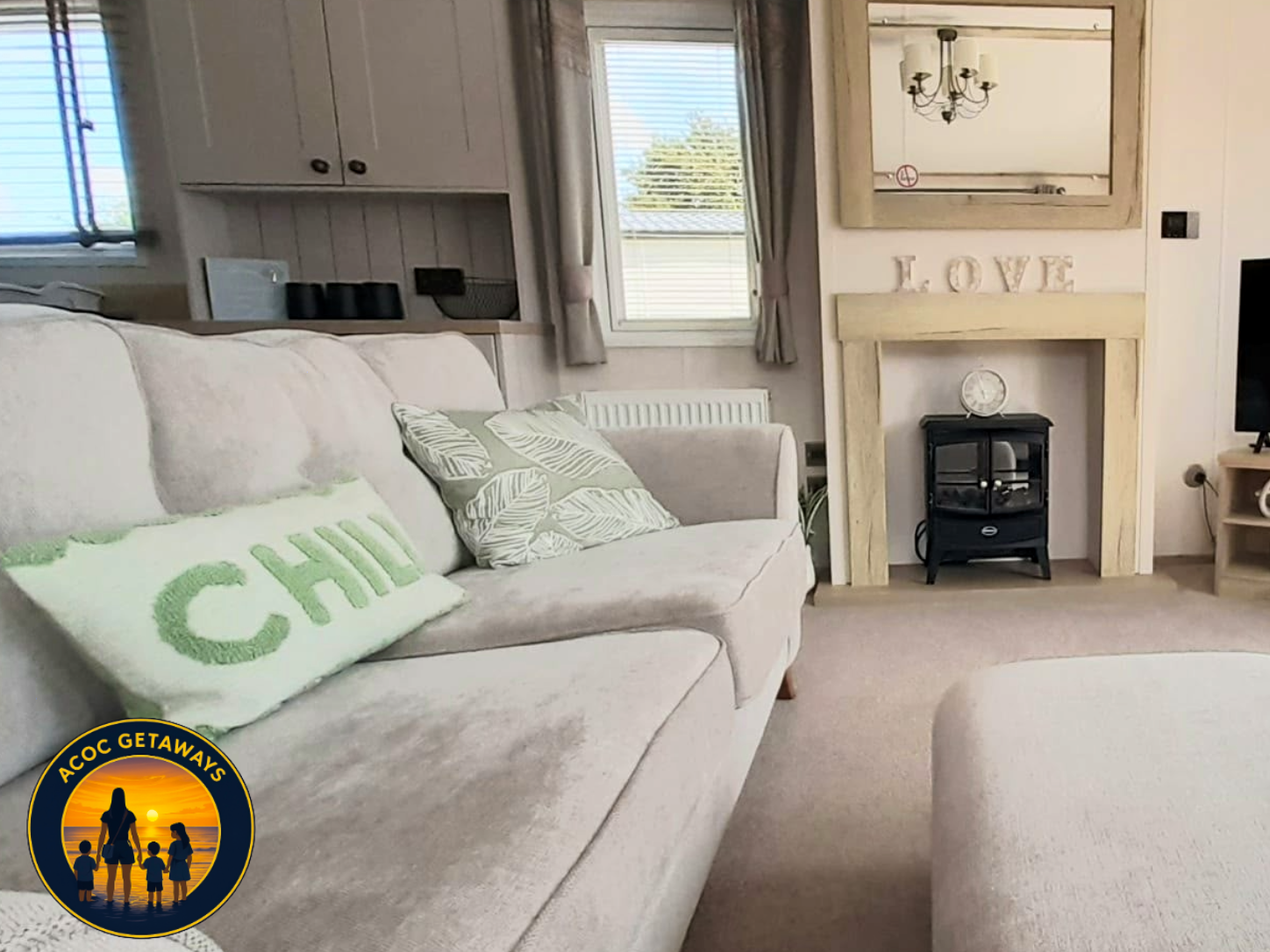 A cozy living room with a beige sofa with pillows, a fireplace with a mirror above, a window with curtains, a chandelier, and a fire stove.