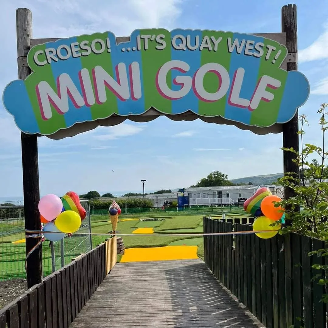 Entrance to Mini Golf course with colorful sign reading "Croeso!... It's Quay West! Mini Golf" and balloons decorated around the entrance.