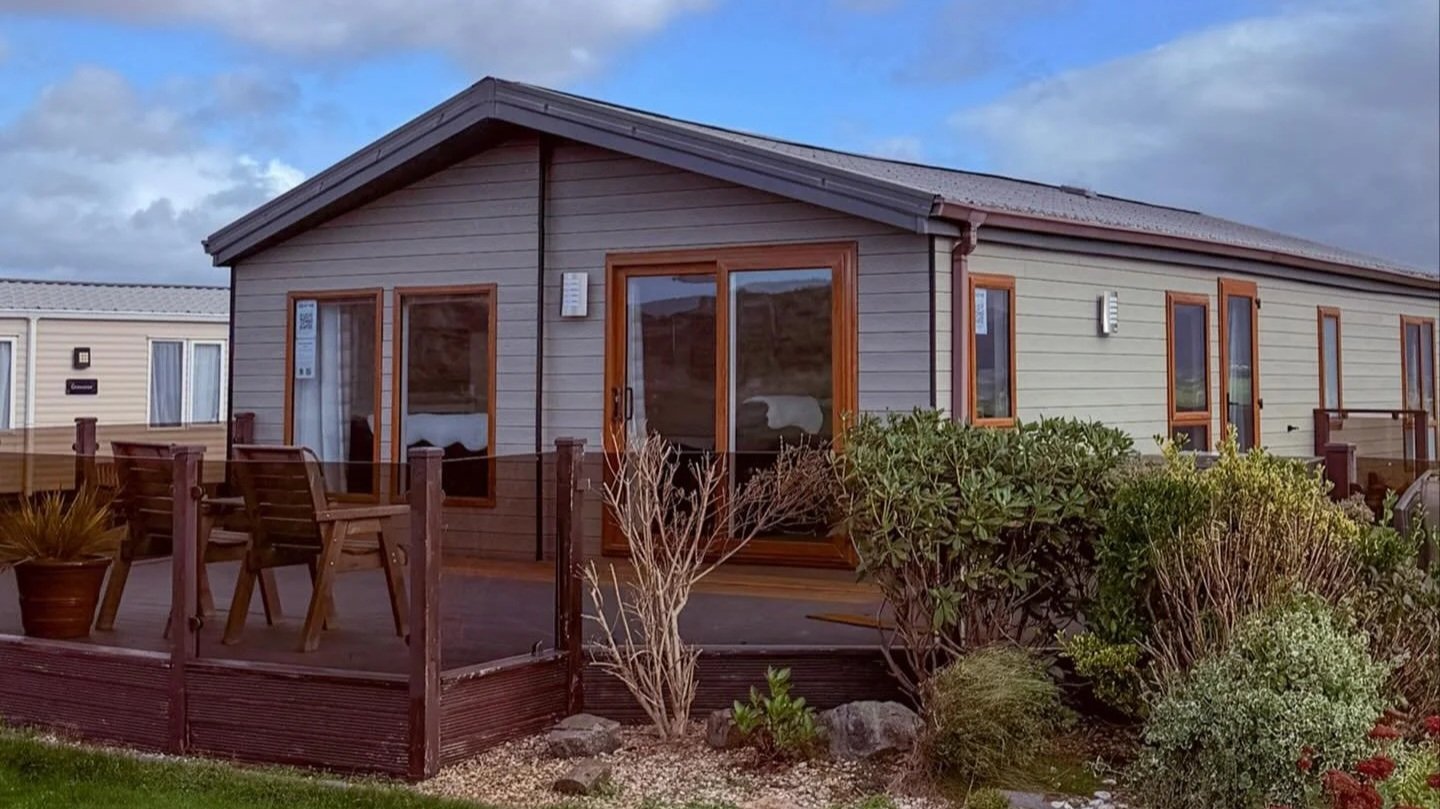 Exterior view of a house with wooden siding, large windows, sliding glass door, small deck with chairs and planters, surrounded by bushes and rocks under a partly cloudy sky.