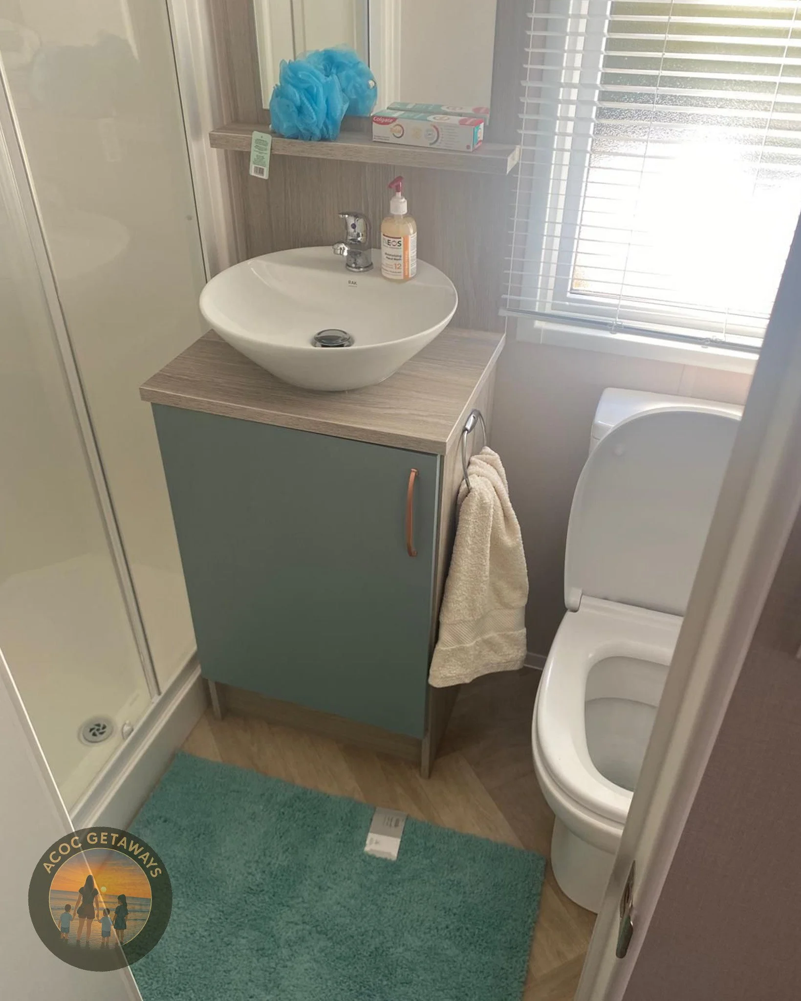 Small bathroom with a corner sink, a blue cabinet, a toilet, a shower with a glass door, a window with blinds, and a bath mat. Items on the shelf include a blue shower pouf, a box of toothpaste, and a bottle of hand sanitizer.