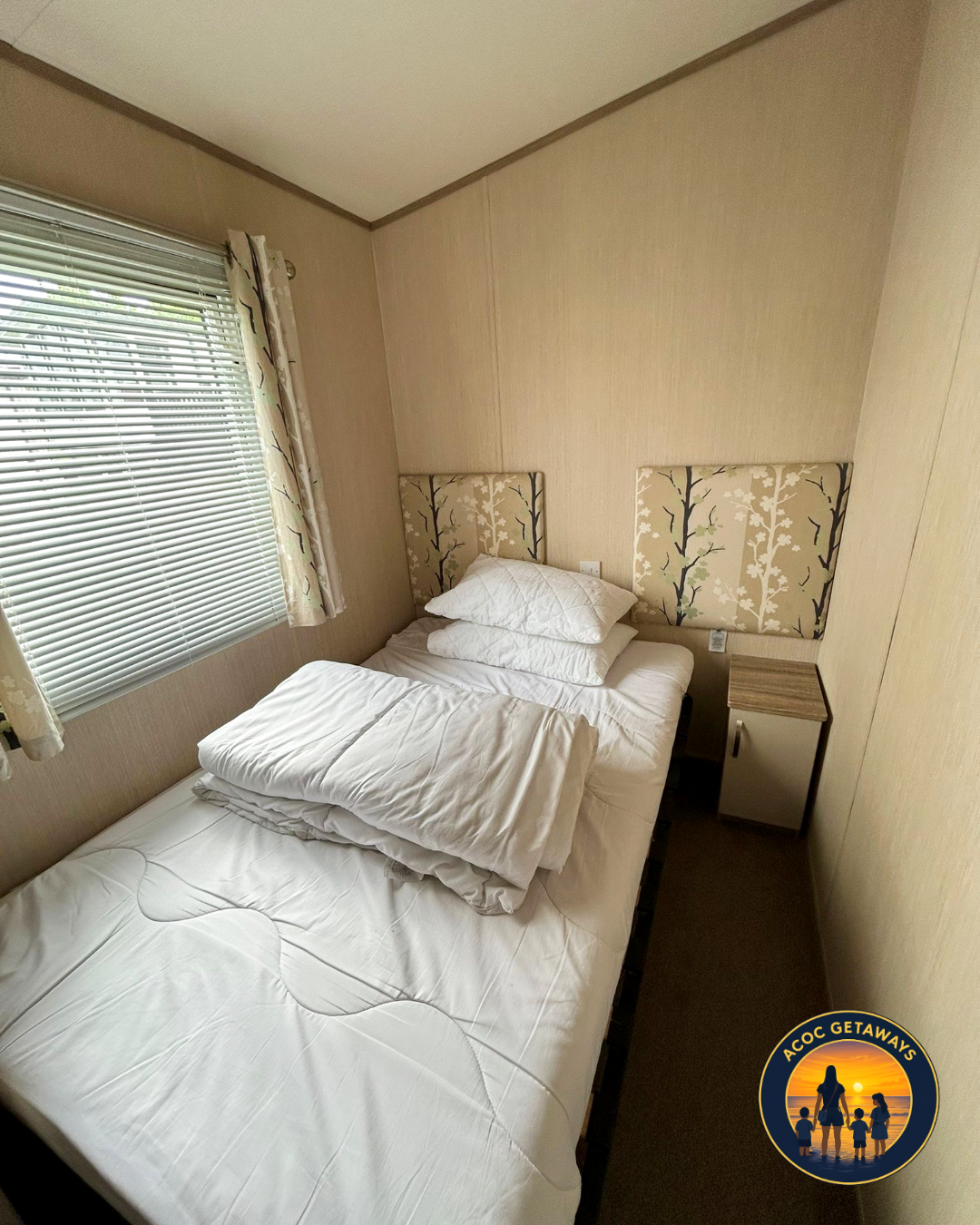 A small bedroom with a bed, pillows, and bedding, next to a window with blinds and floral curtains, and a nightstand with a cabinet.