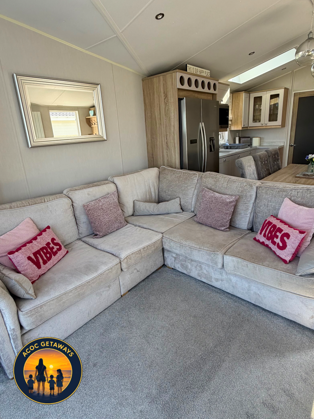 A cozy living room with a beige sectional sofa, decorated with pink and purple pillows, including two with 'VIBES' written on them. A large mirror hangs on the wall above the sofa. The background shows a kitchen area with a stainless steel refrigerat