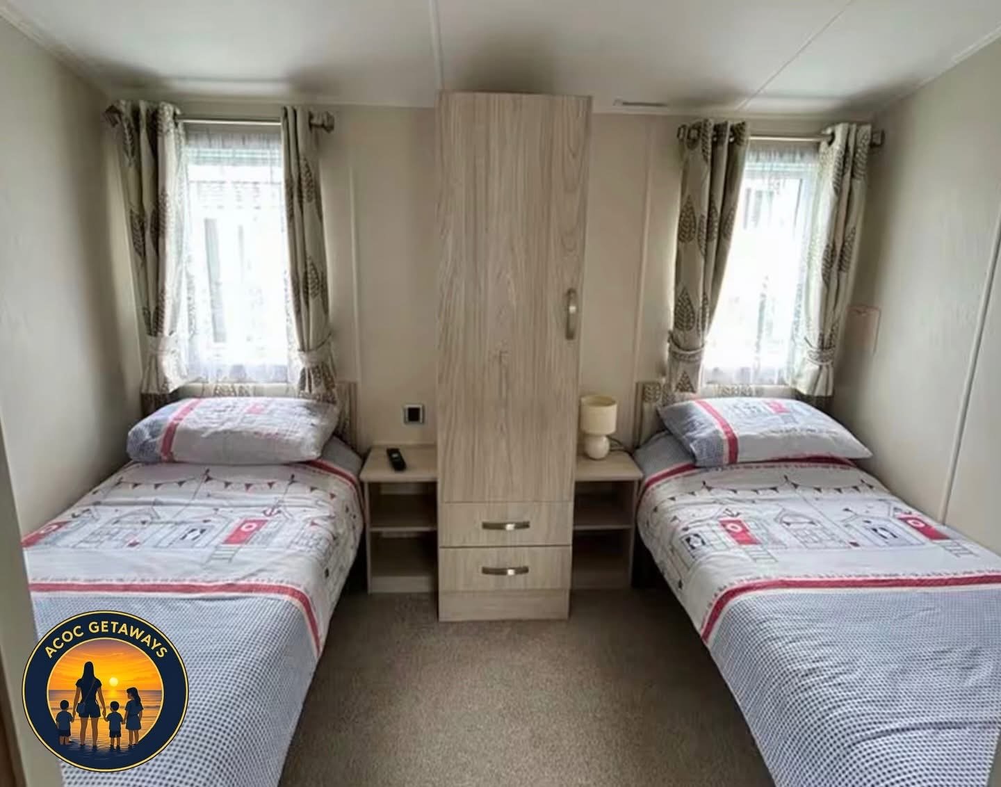 A bedroom with two twin beds, each with patterned bedding and pillows, separated by a nightstand with a lamp and a remote, in front of two windows with patterned curtains.