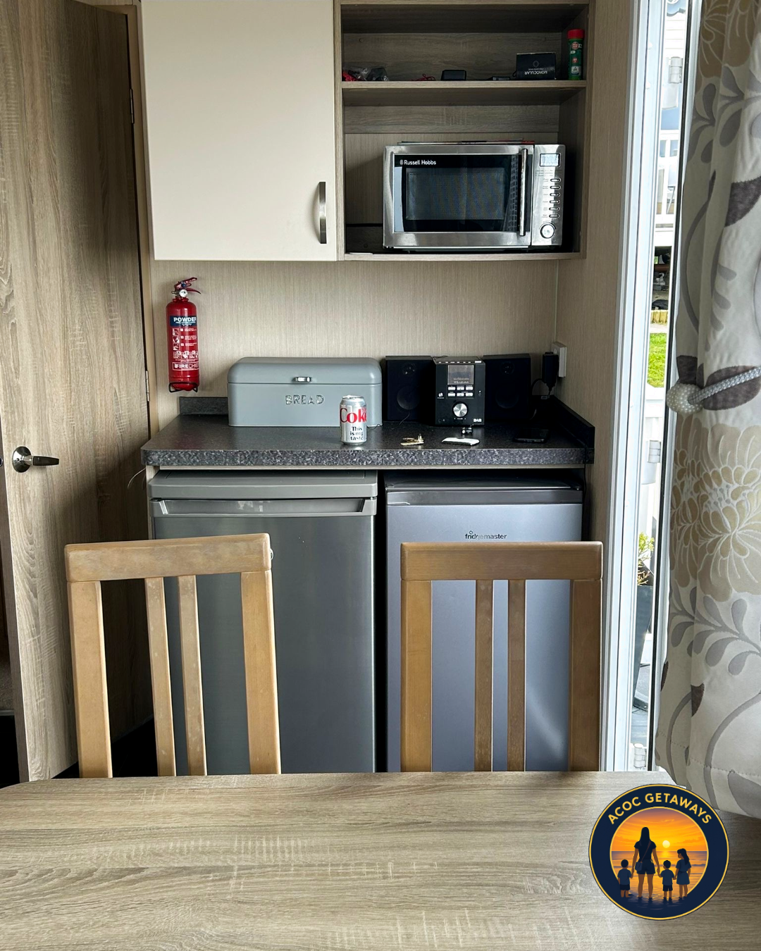 Kitchen area with two mini refrigerators, counter with a bread box, soda can, and key, microwave, stereo system, fire extinguisher, and chairs at a wooden table. An open doorway reveals part of a balcony.
