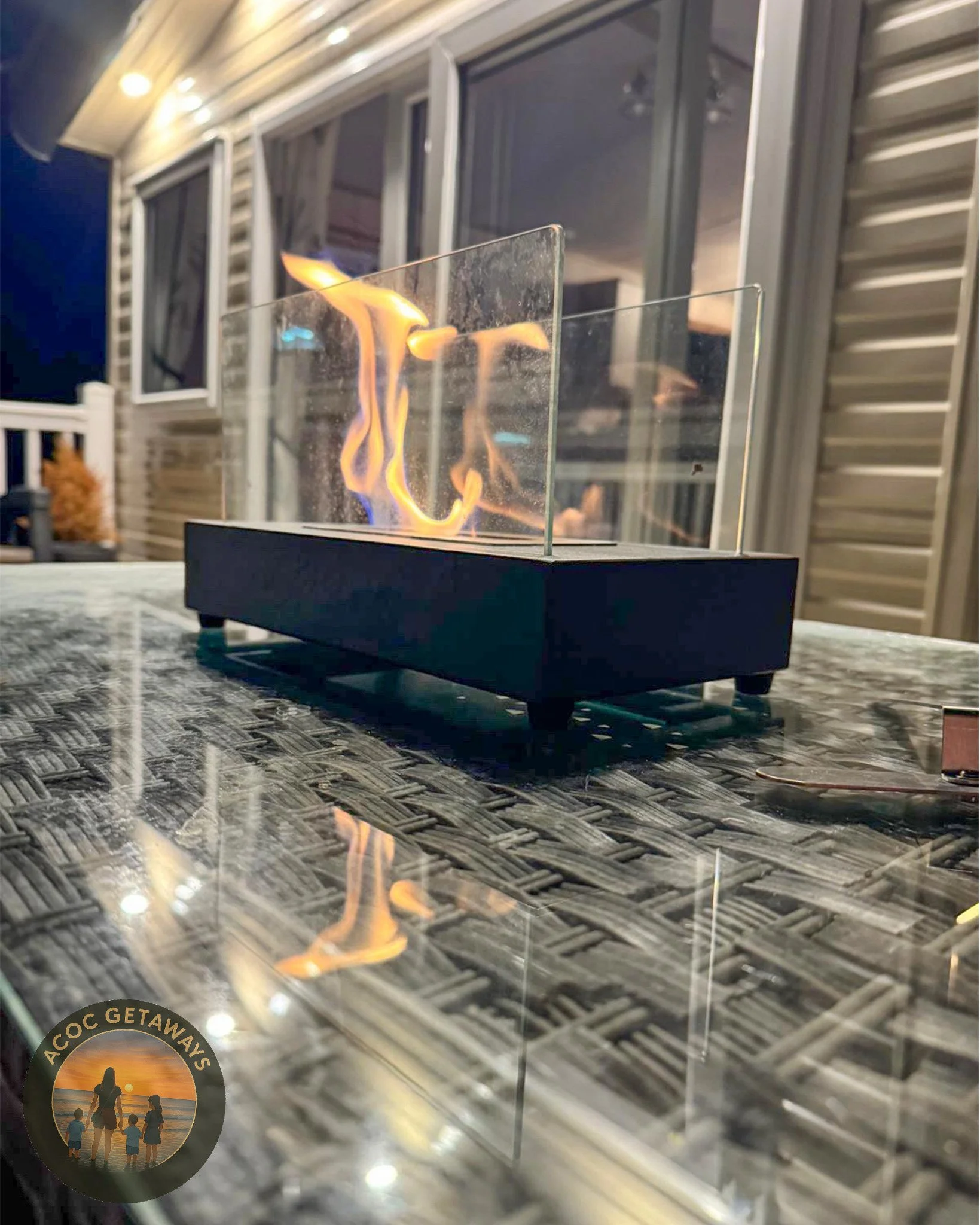 A tabletop fireplace with flames behind a glass barrier on a woven-texture table outside at night.