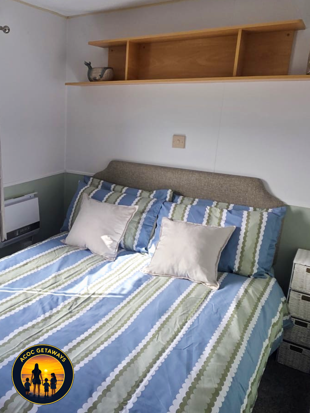 A bedroom with a bed covered in a blue, green, and white striped bedspread. There are two white pillows and a brown headboard. Above the bed is a wooden shelf with a ceramic deer figurine. The room has white walls with a lower green trim, and a wicke