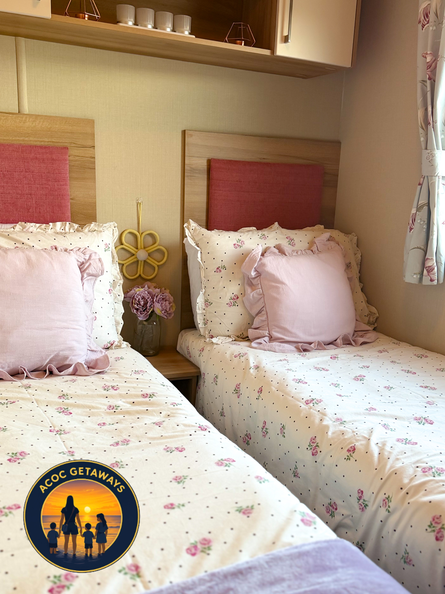 Two beds with pink headboards, floral bedspreads, and pink pillows in a cozy room. A nightstand with a glass jar of purple flowers, decorative wall hanging, and shelves with cups and decor are visible.