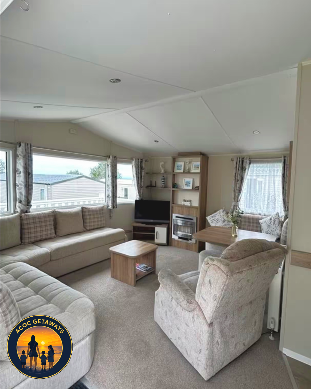 Cozy living room with beige couches, floral curtains, a wooden coffee table, and a small dining area with a table and armchair. There is a TV, fireplace, and bookshelf, with windows showing a mobile home park outside.
