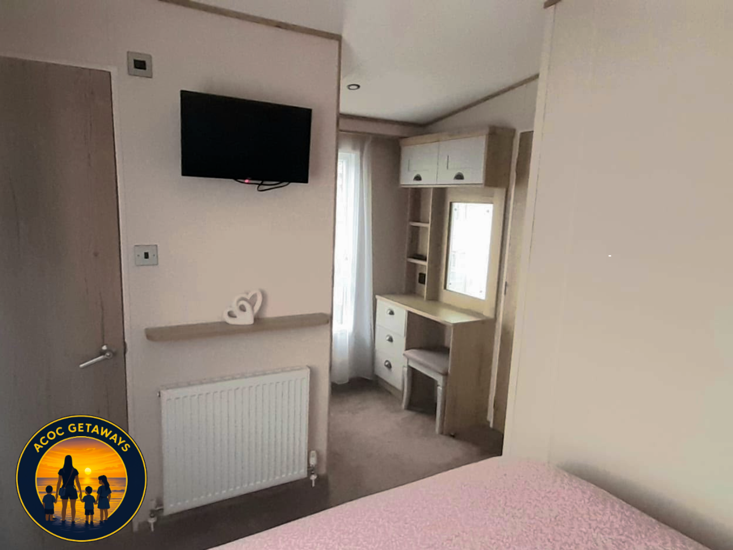 Interior of a bedroom with a wall-mounted TV, a white built-in desk with drawers, a window with curtains, and a bed with pink bedding.