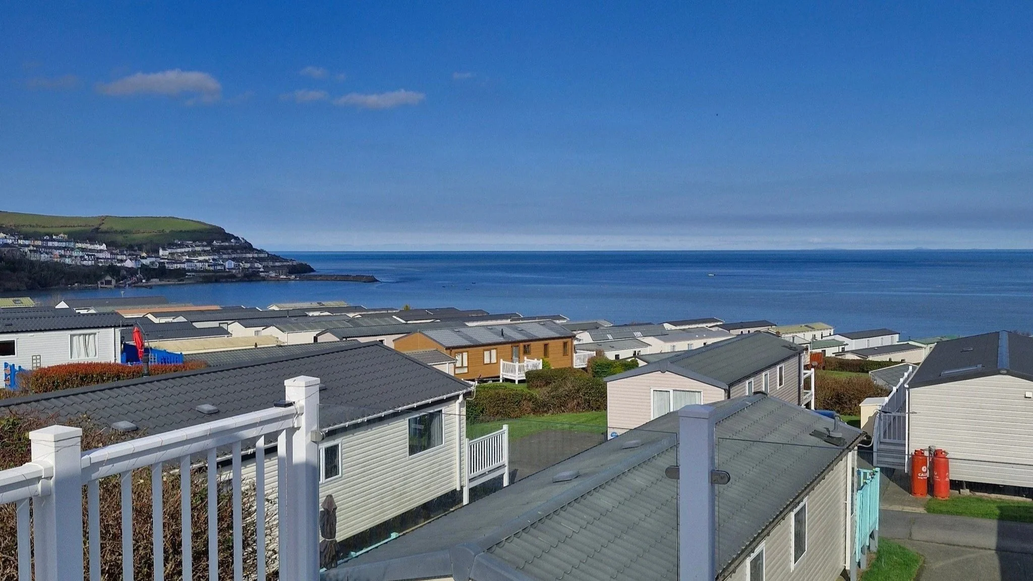 View of a coastal neighborhood with houses on a hillside overlooking the ocean, with a hill in the background under a partly cloudy sky.