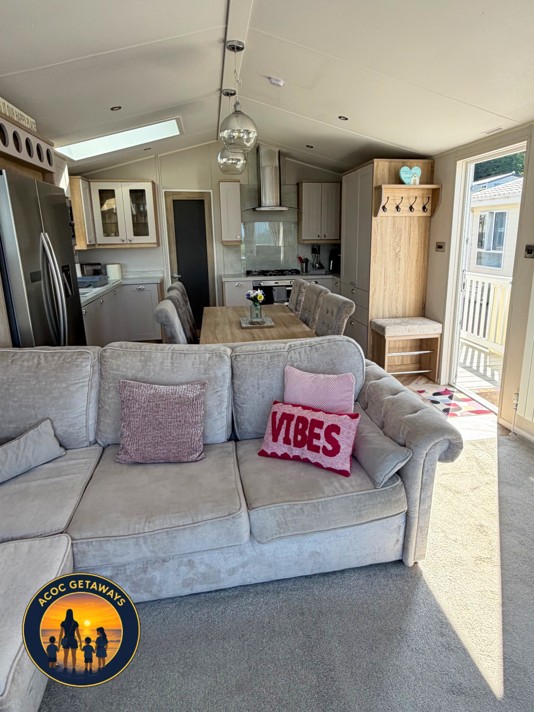 Open-plan living room and kitchen with a beige sectional sofa, decorative pillows, a wooden dining table with chairs, kitchen appliances, and large windows letting in natural light.