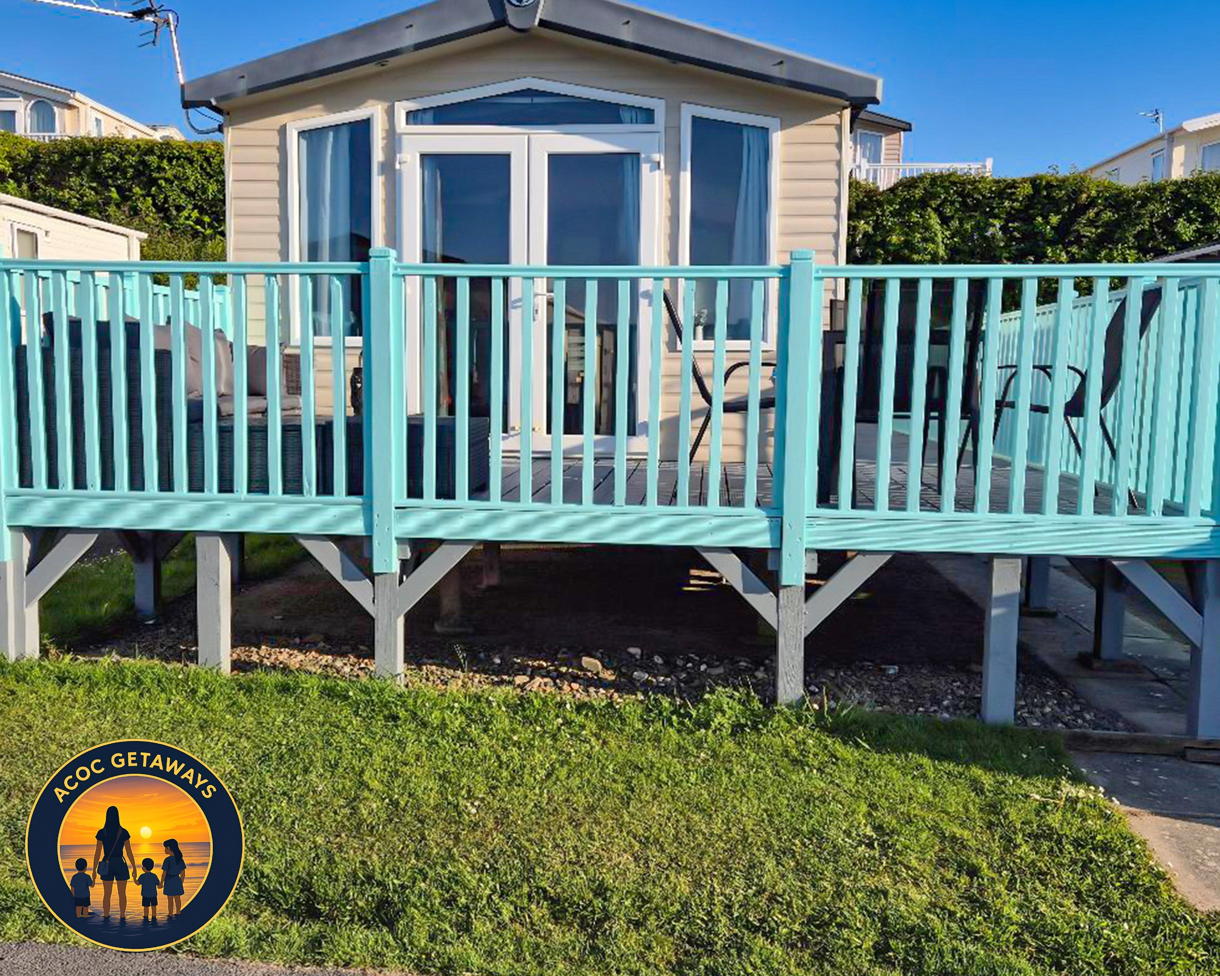 A house with a light beige exterior, large glass doors, and a turquoise balcony railing. There is outdoor furniture on the deck and a grassy area in the foreground. The sky is clear and blue.