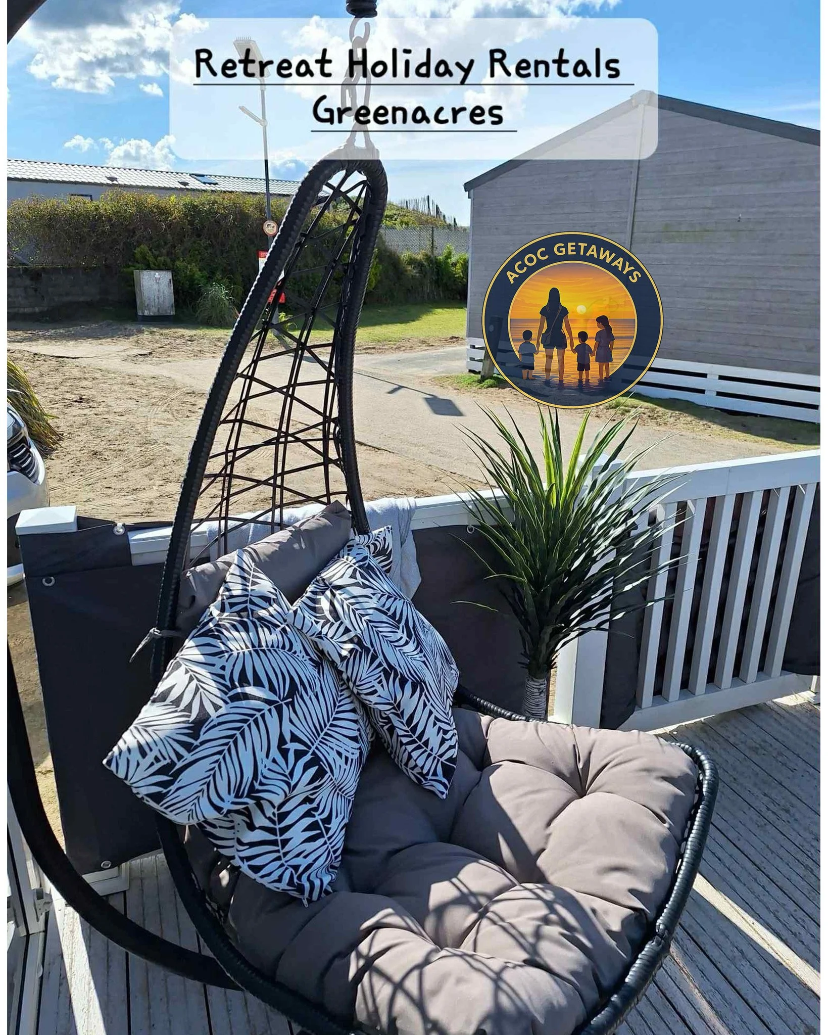 A hanging outdoor swing chair with beige cushions and two black and white leaf patterned pillows, situated on a deck next to a white railing. A potted green plant and a background with a dirt lot, a grey house, and a blue sky with some clouds. There 
