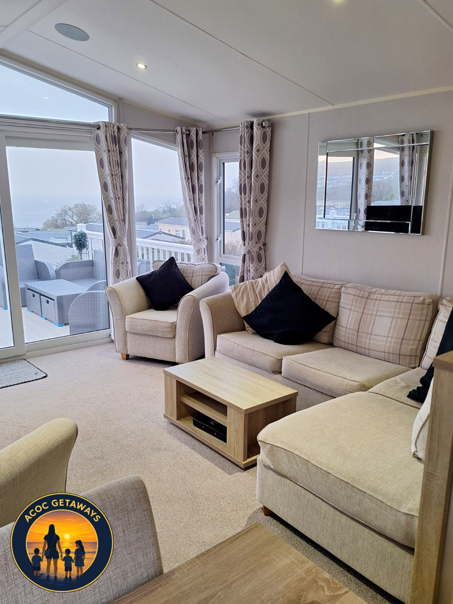 Living room with beige couches, black pillows, patterned curtains, glass sliding door leading to a balcony with outdoor furniture, large window showing overcast sky, wall mirror, light wood coffee table, and beige carpeted floor.