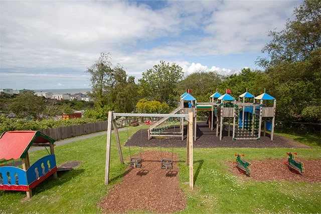 Playground with swings, slides, and spring riders in a grassy area with trees and a sky with clouds.