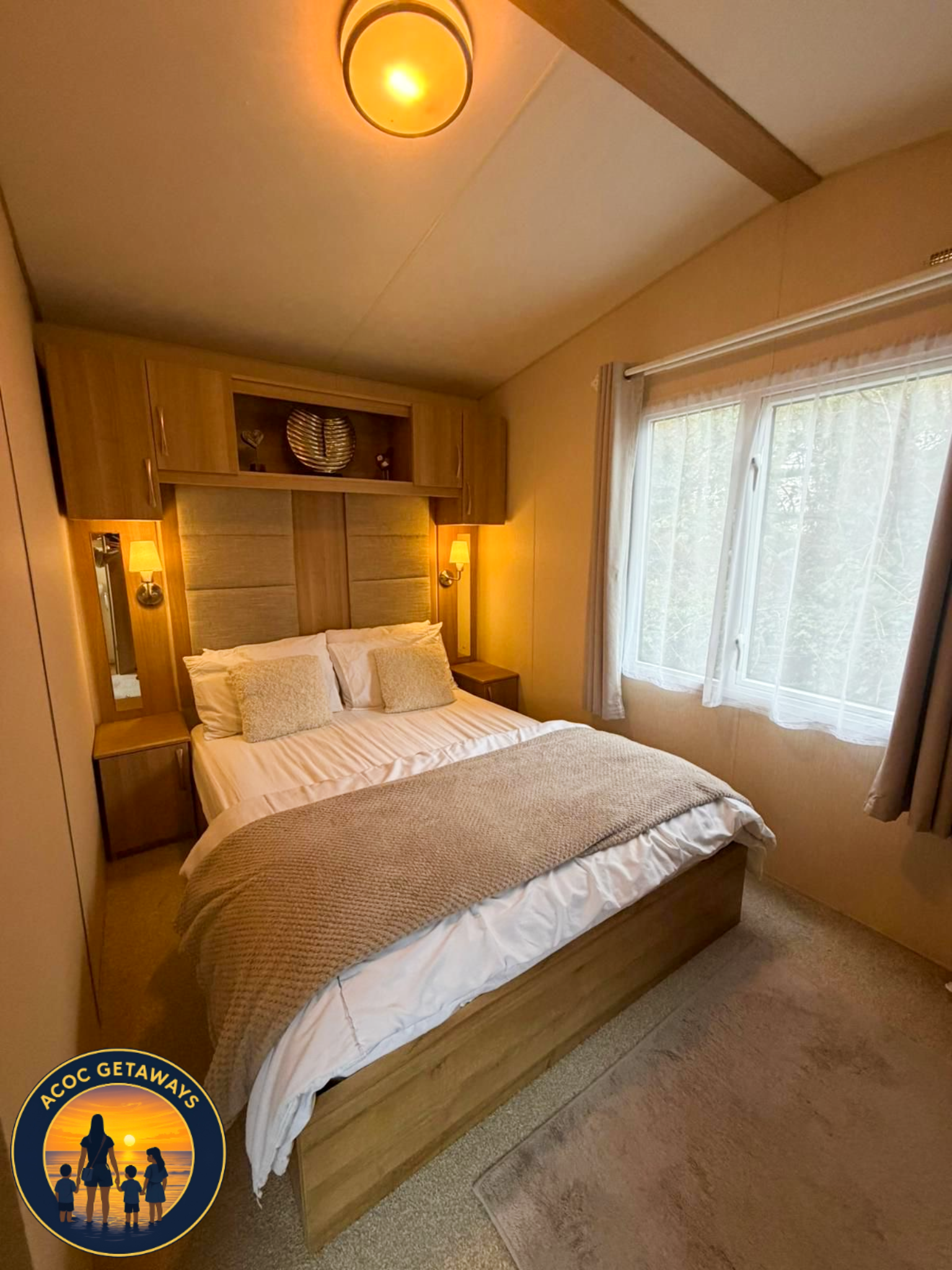 A cozy bedroom with a large bed, beige bedding, and matching pillows. There are two nightstands with lamps on each side of the bed, and a window with sheer curtains letting in natural light. A ceiling light is also visible.