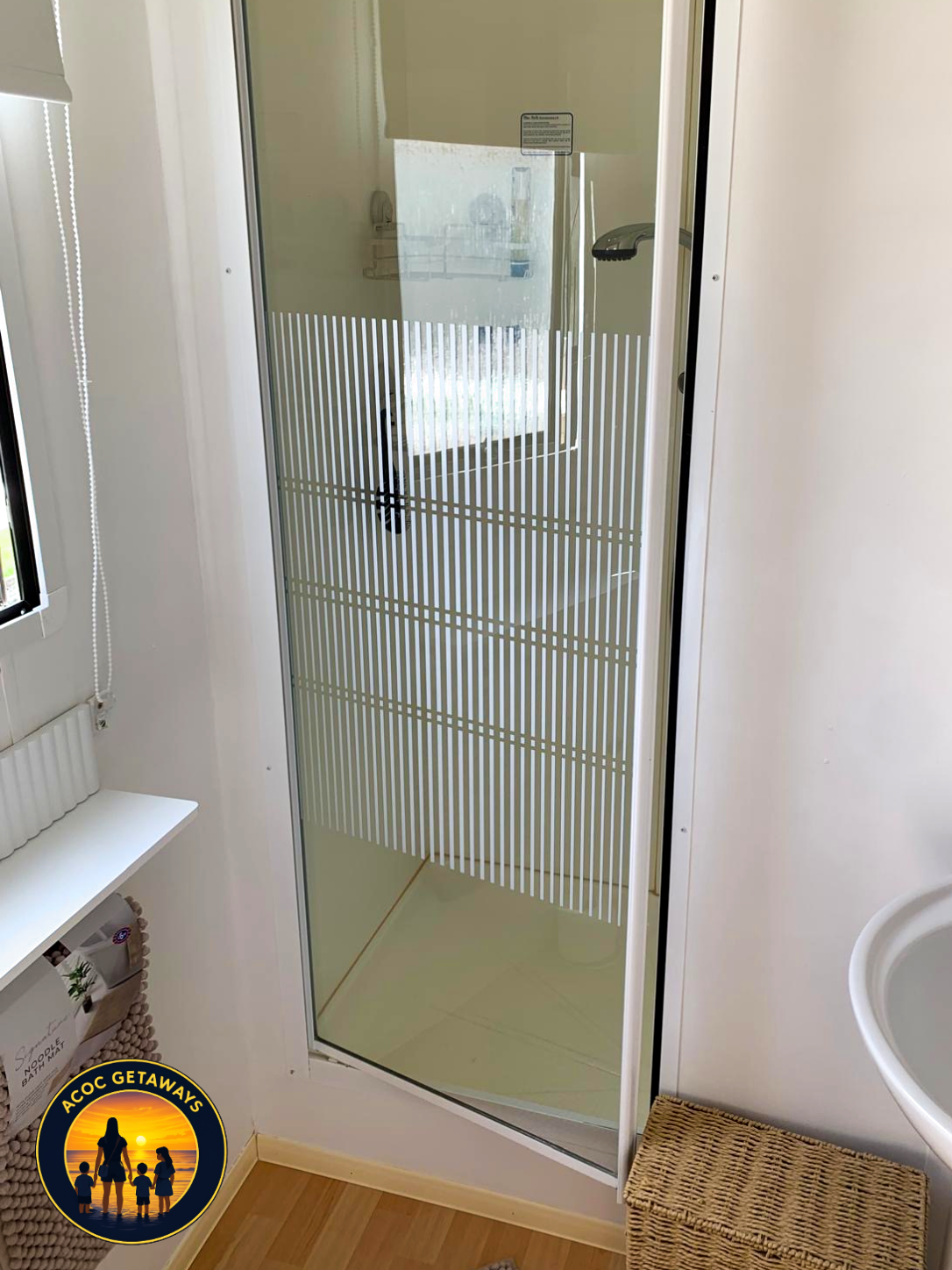 Interior of a small bathroom with a corner shower with a frosted glass door and striped design, a window, a white shelf with a basket underneath, and a woven mat on the wood floor.
