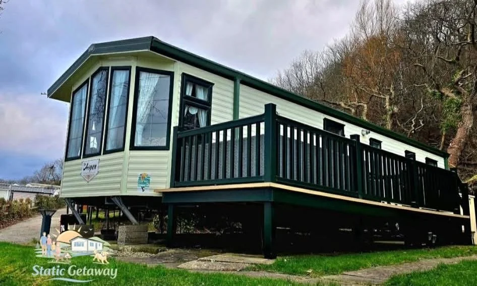 A raised tiny house with a green roof and siding, large windows, and a black railing porch, situated on a grassy area with trees in the background.