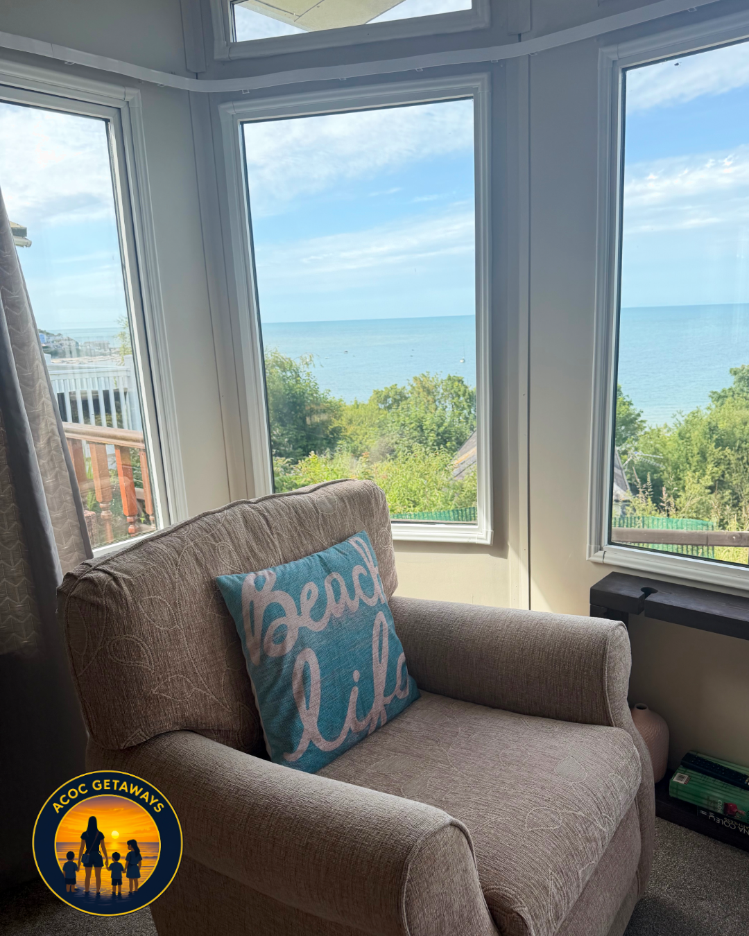 Cozy living room with a beige armchair that has a pillow reading 'Beach Life,' large windows showing a view of green trees, a blue sea, and a partly cloudy sky.