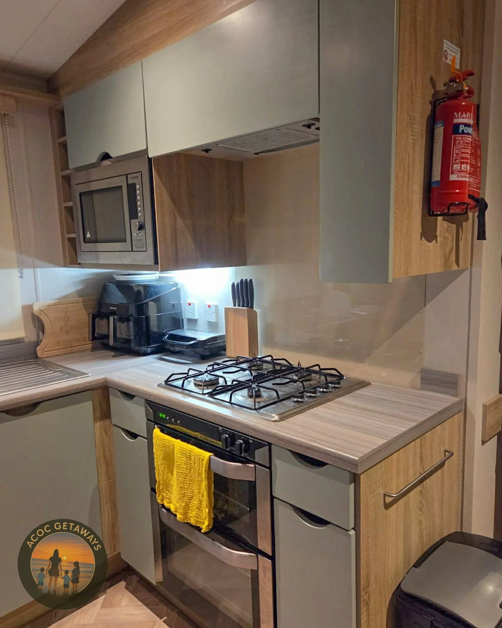 Compact kitchen with microwave, coffee maker, knives, stove, oven, yellow towel, fire extinguisher, and ACOC Getaways logo.