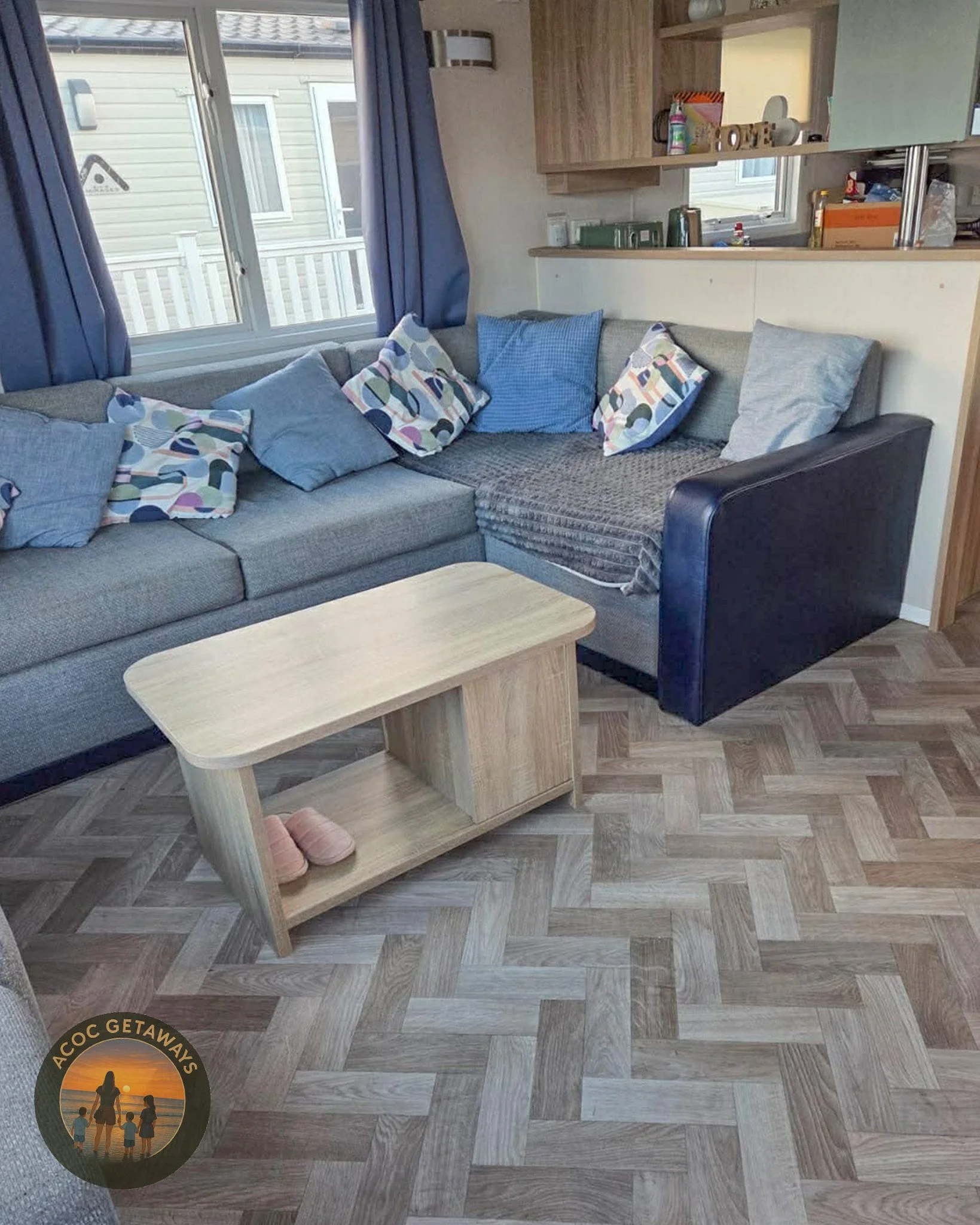 Living room with a large sectional sofa, multiple colorful and blue pillows, a light wood coffee table, and two windows with blue curtains. There are various items on the shelves behind the sofa, and a sticker with a sunset and the text "ACOC GETAWAYS" in the bottom left corner.