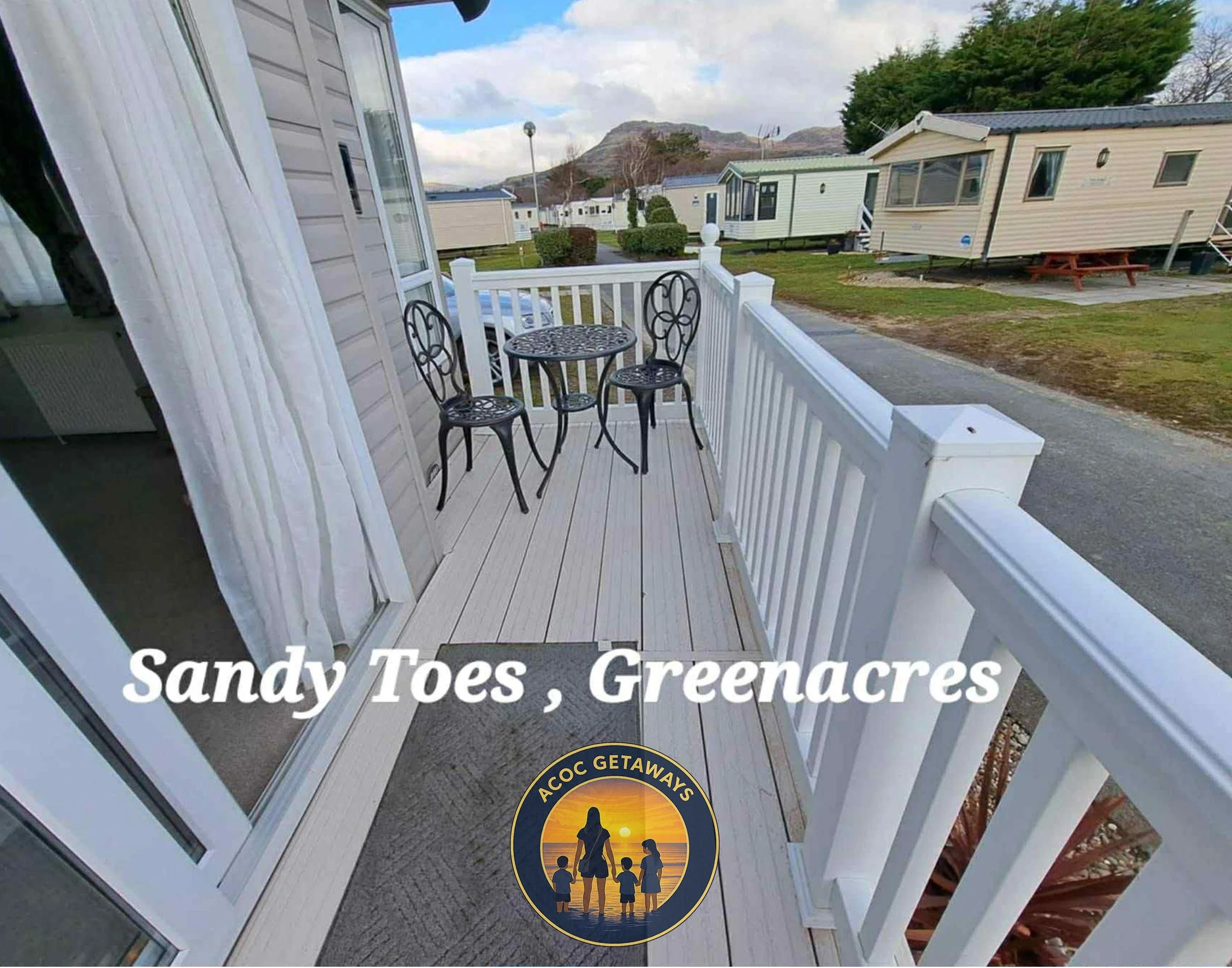 View of a small outdoor balcony with a round table and two chairs, overlooking a row of mobile homes in a campground with mountains in the background.