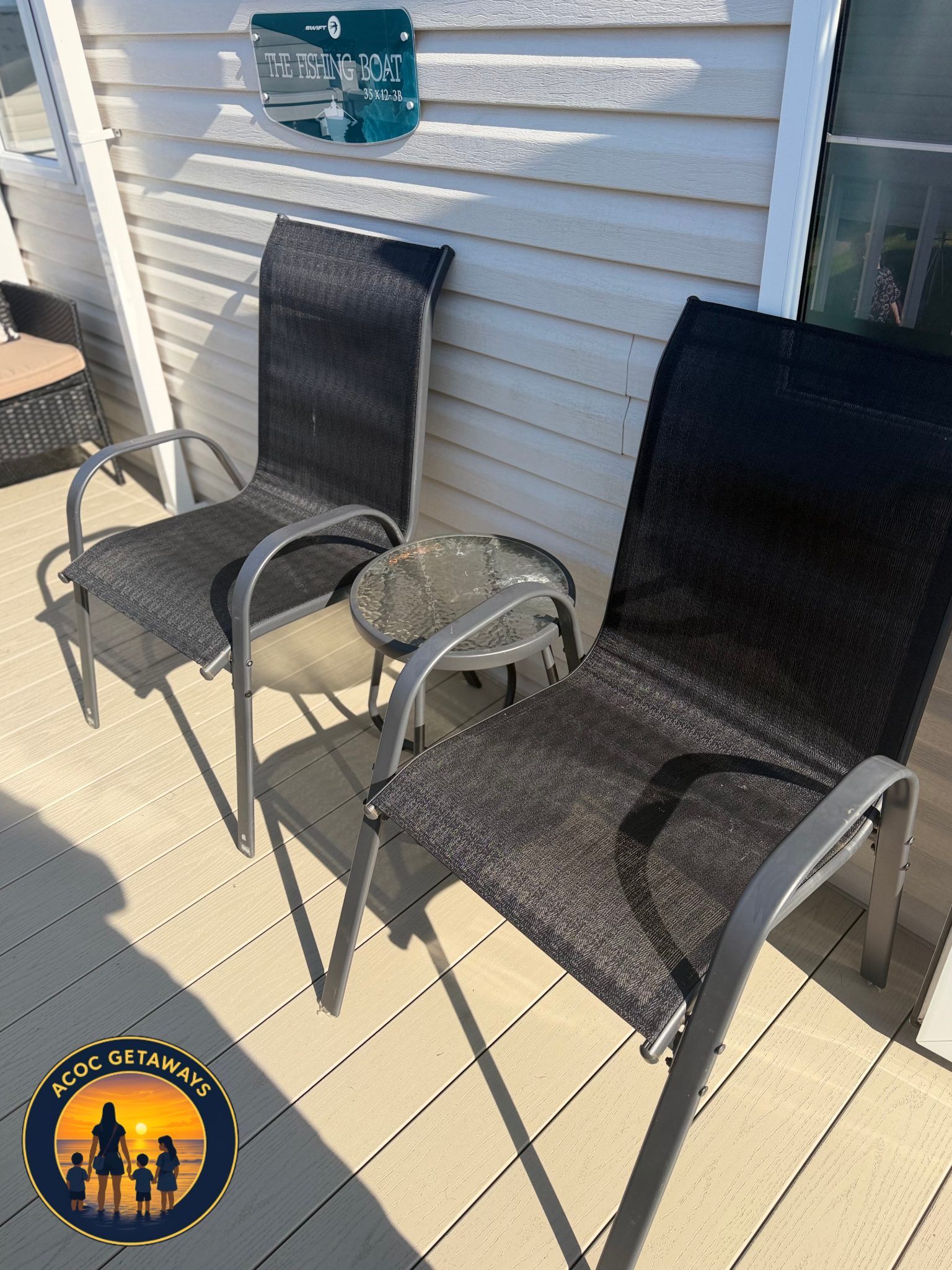 A patio or deck area with two black mesh chairs, a small round glass-topped table, and white vinyl siding on the house wall. A sign on the wall reads 'The Fishing Boat'. There is a sliding glass door and a partial view of a person in the reflection. 