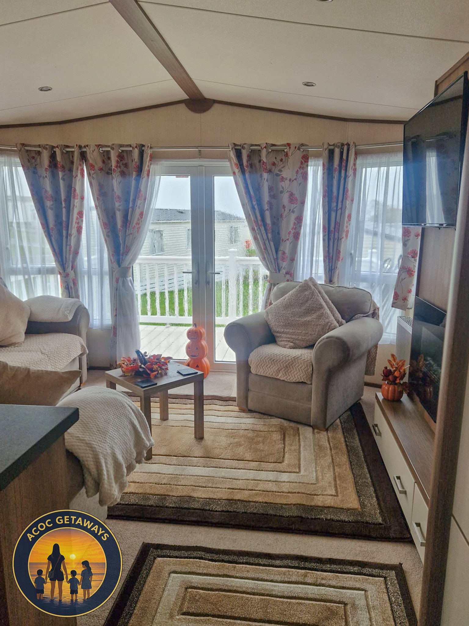 A cozy living room with large windows covered by floral curtains, beige armchairs, a small coffee table with colorful decorations, and a TV cabinet. There are outdoor views of a deck and houses beyond.