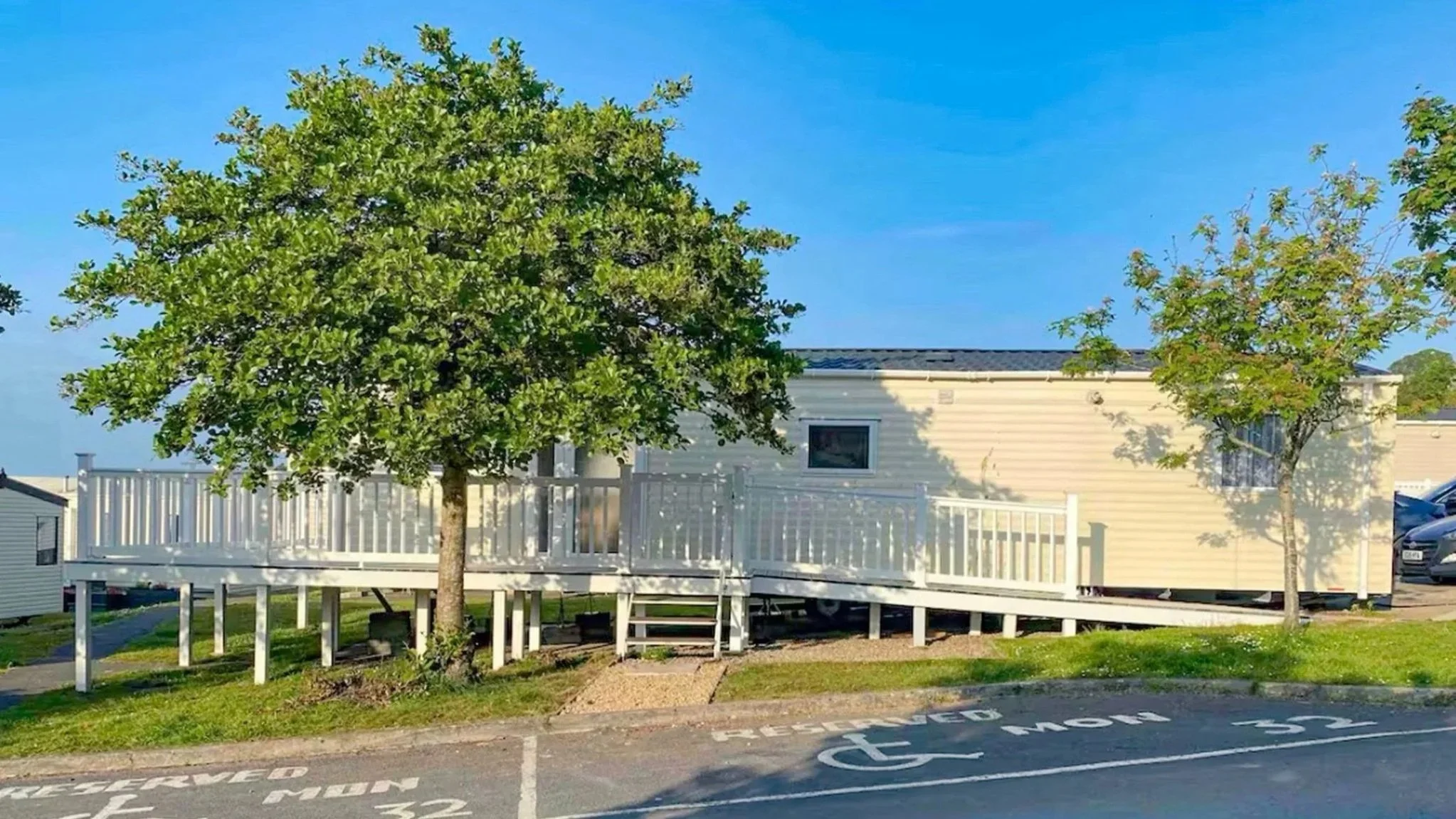 A mobile home with beige exterior siding, surrounded by a white railing deck, two trees, a parking lot with marked spaces including a wheelchair accessible spot, and a blue sky overhead.