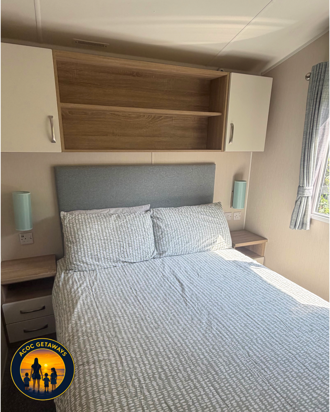 A bedroom with a double bed, a gray headboard, patterned bedding, two bedside tables with small drawers, and wall-mounted lamps. There is a wooden wall-mounted shelf above the bed and a window with curtains to the right.