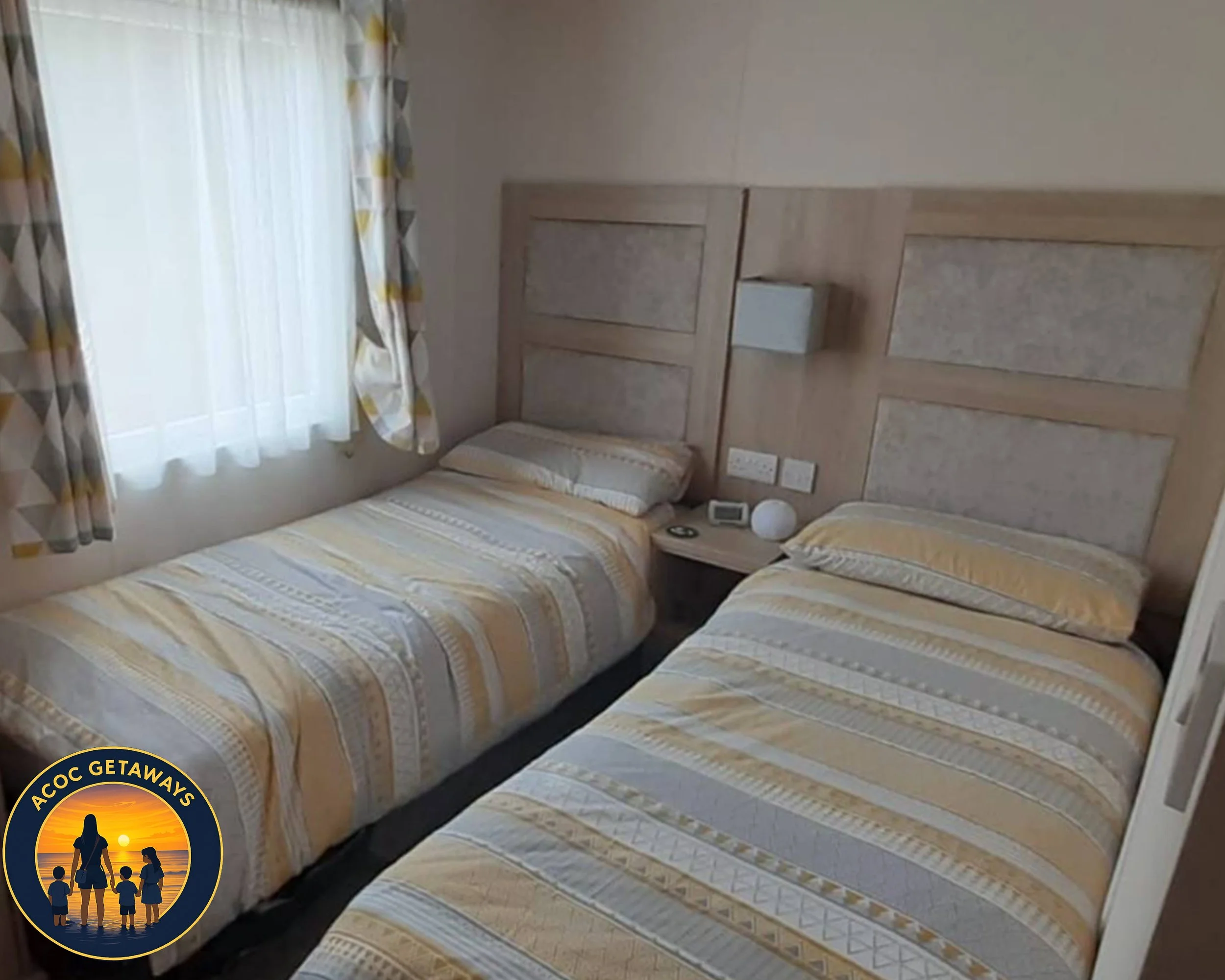 Two single beds with matching yellow and white striped bedspreads in a hotel room with a window dressed with patterned curtains, a nightstand with a clock and a lamp nearby the beds, and wall outlets above the nightstand.