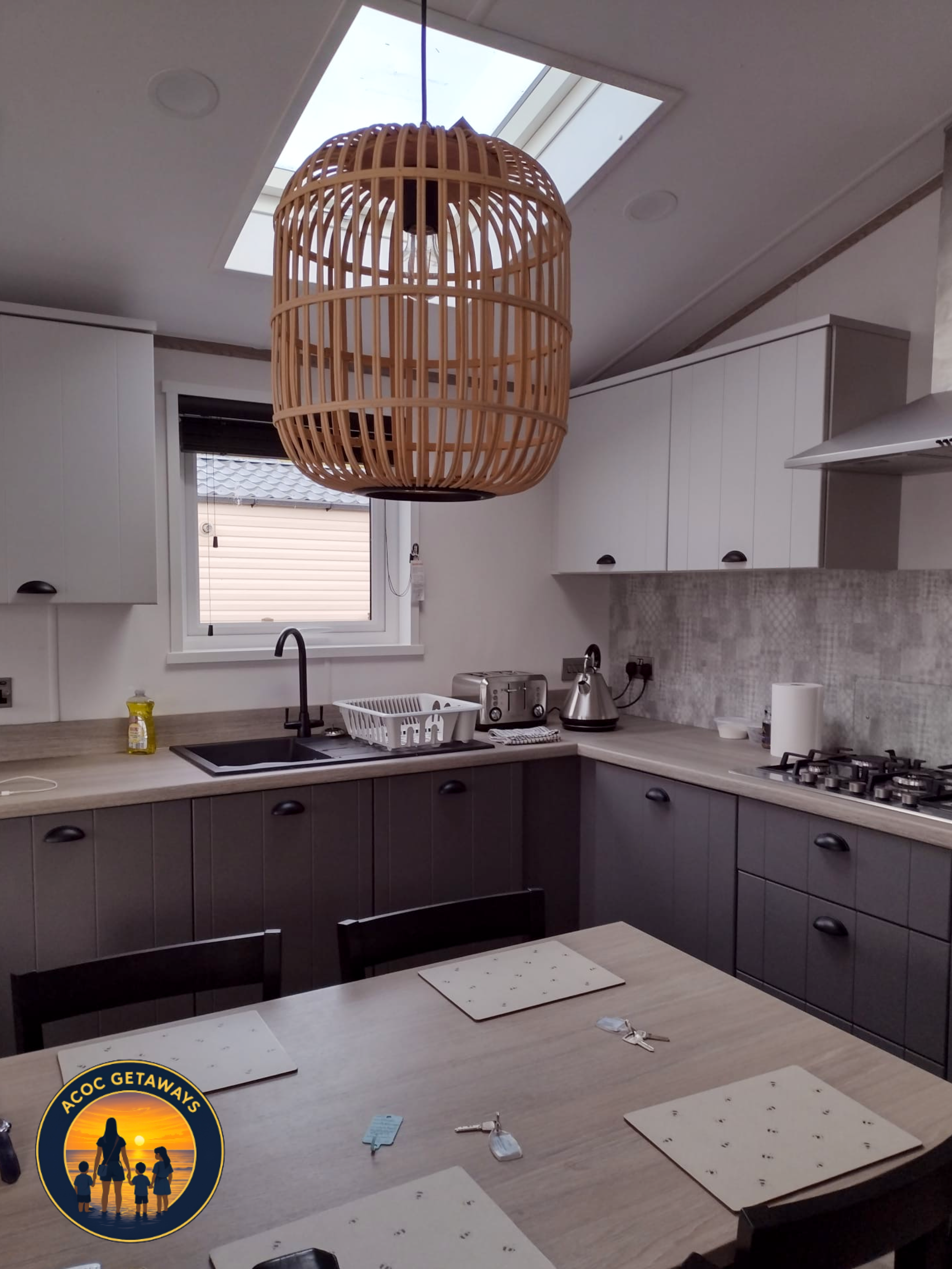 A kitchen with white cabinets, a black sink, a wooden dining table with placemats, a kettle, a toaster, a window, and a wooden pendant lamp hanging from the ceiling.