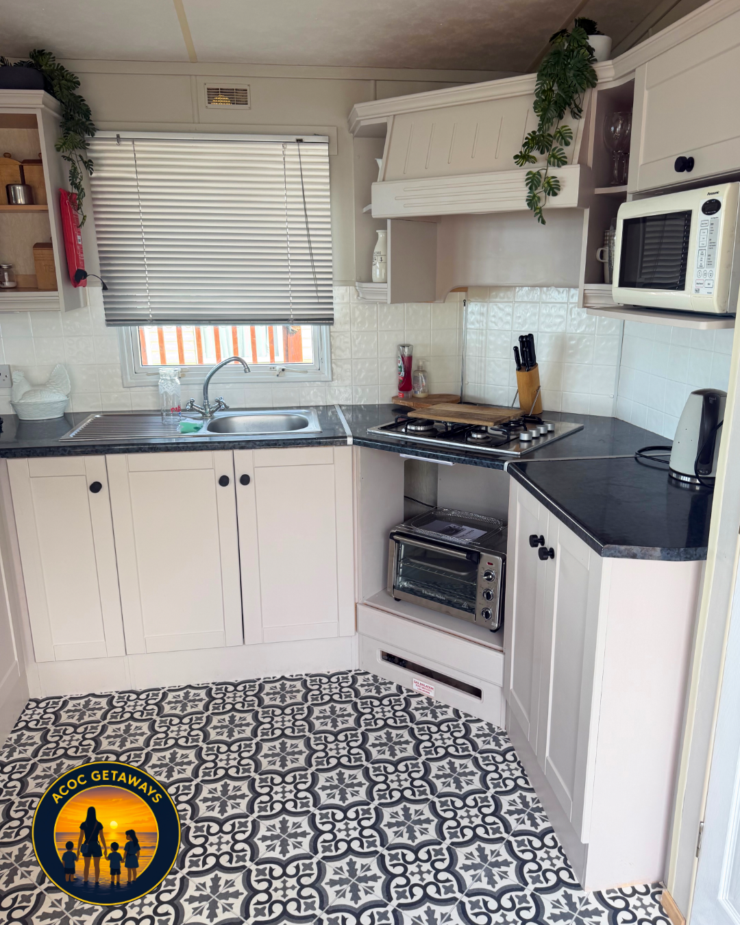 A cozy kitchen with white cabinets, a black countertop, a window with blinds, a sink, a stove with knives on a wooden cutting board, a microwave, a toaster oven, a kettle, and decorative green plants on top of the cabinets. The floor has patterned bl