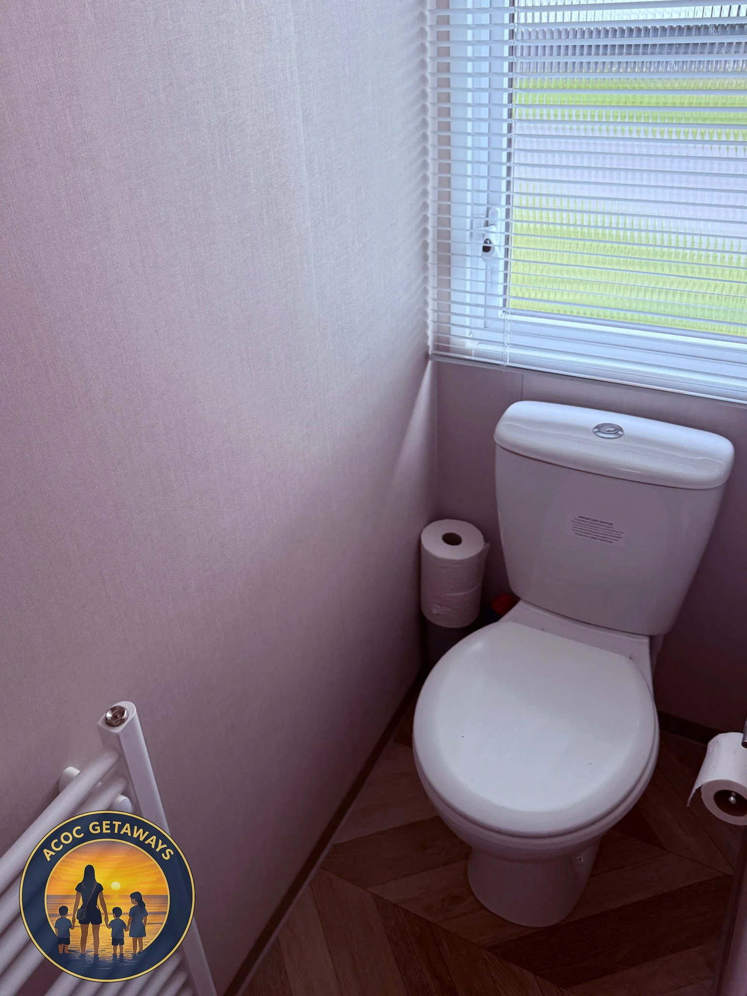 A small bathroom with a white toilet, a roll of toilet paper on the ground, a window with blinds, and a gray wall.