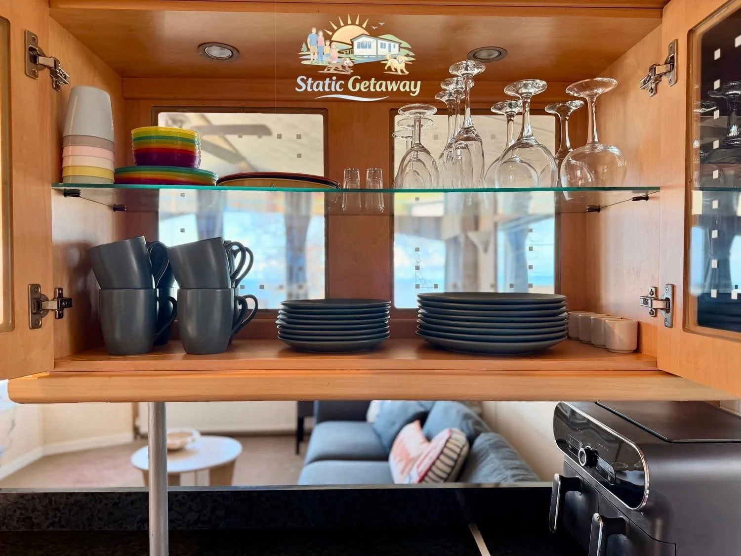 Open wooden cabinet with plates, cups, and glassware inside. Reflection showing a living room with a sofa and window.
