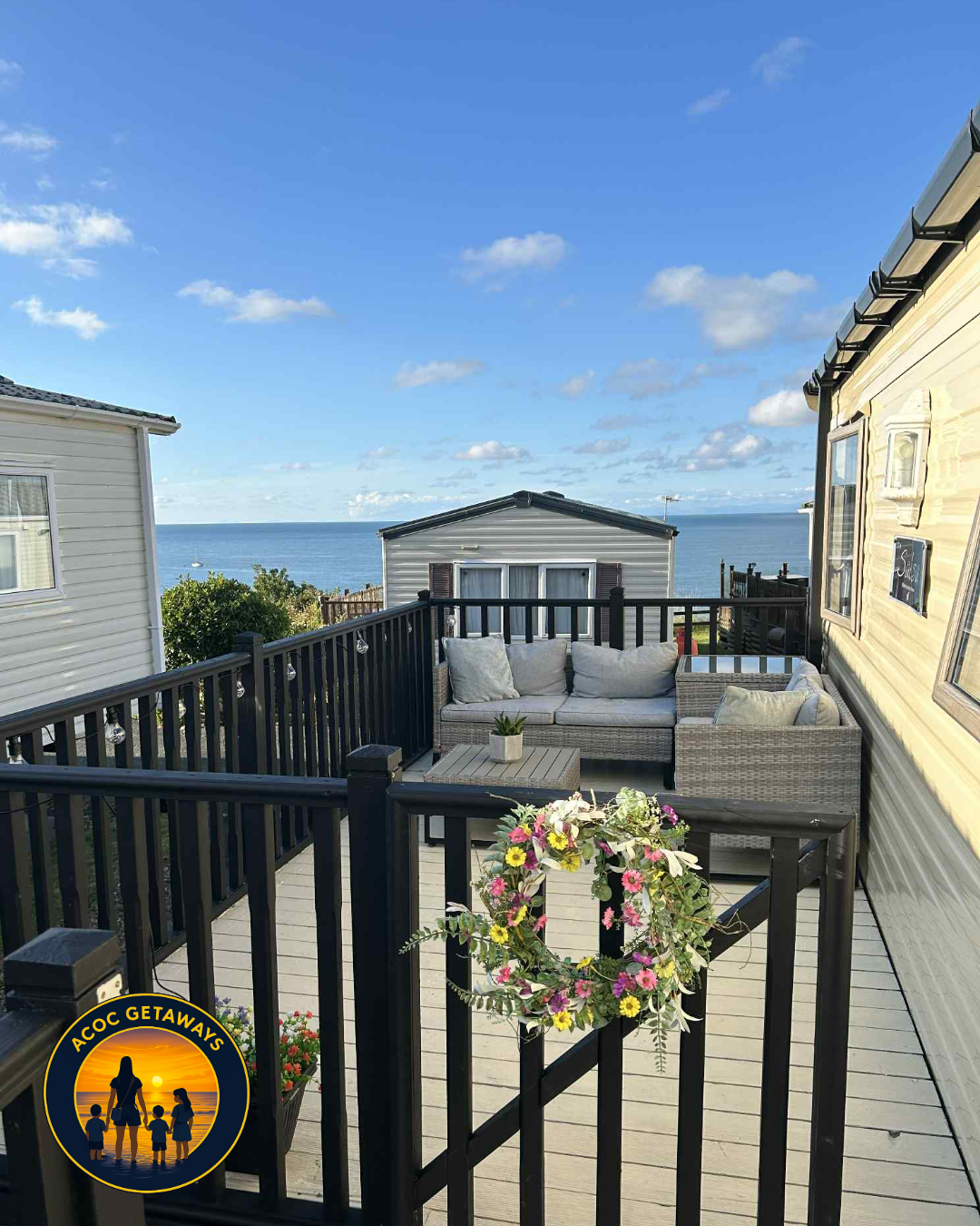 Outdoor wooden deck with black railing, decorated with a floral wreath, overlooking ocean under a blue sky with scattered clouds.