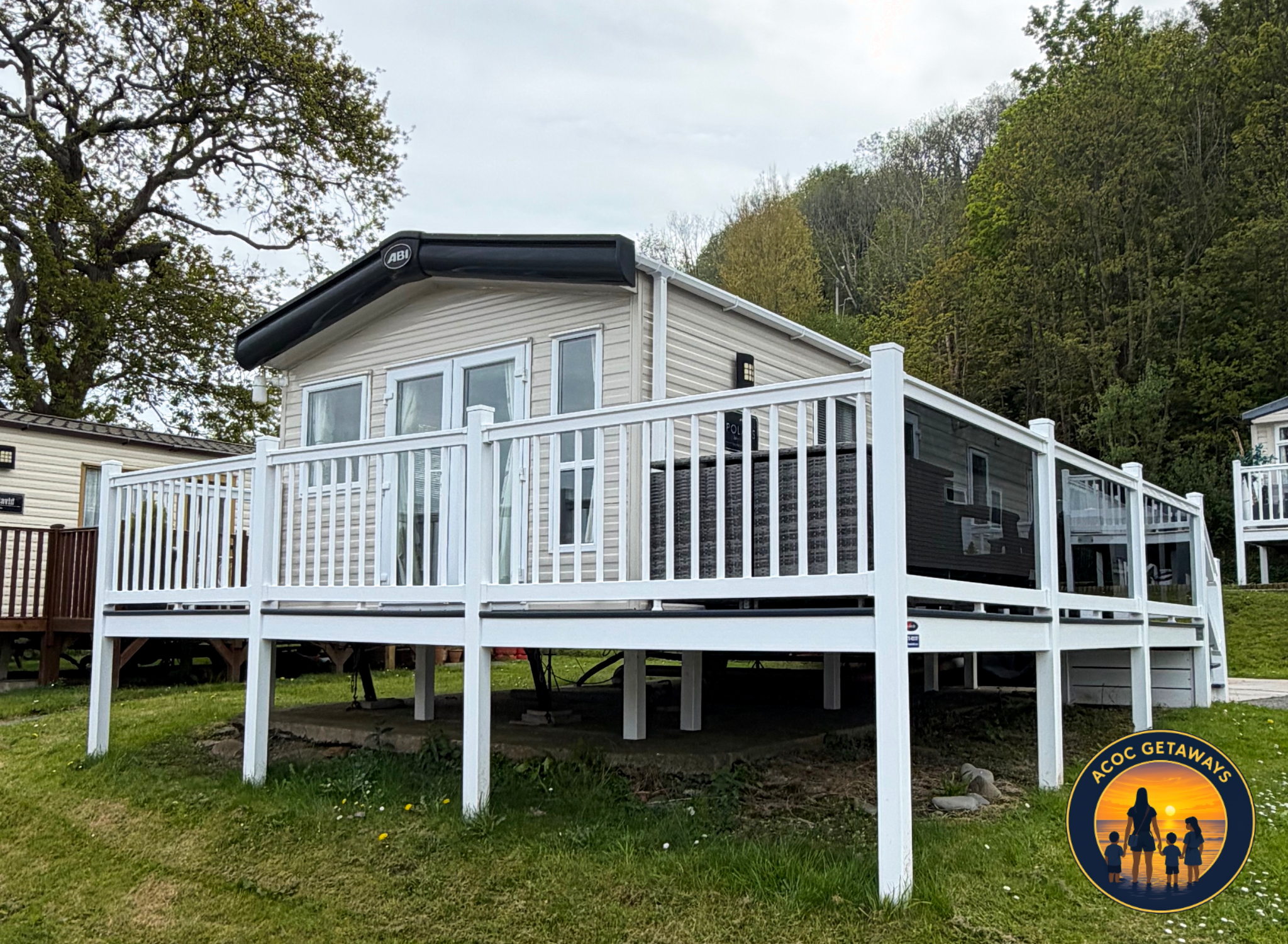 A two-story beige house with white trim and a deck in the backyard. The house has multiple windows, including a sliding door. The deck has white railings and contains outdoor furniture. The house is surrounded by grass, rocks, and trees, with a hills