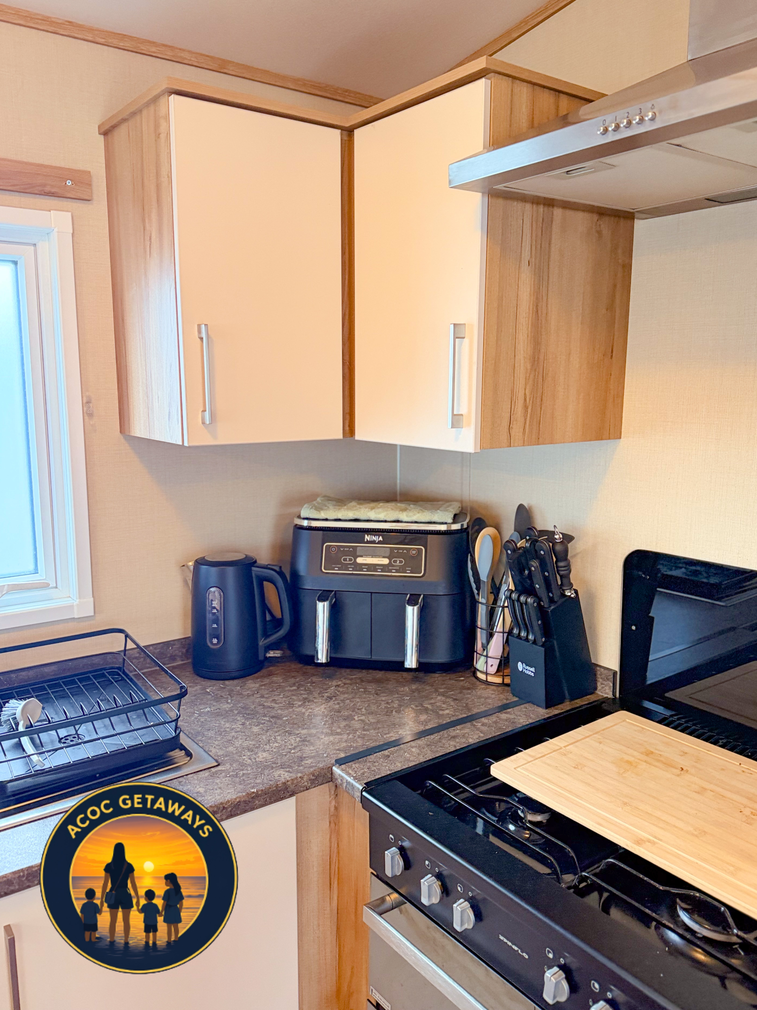 Kitchen countertop with black air fryer, electric kettle, knife block, and dish rack, with wooden cabinets and stove.
