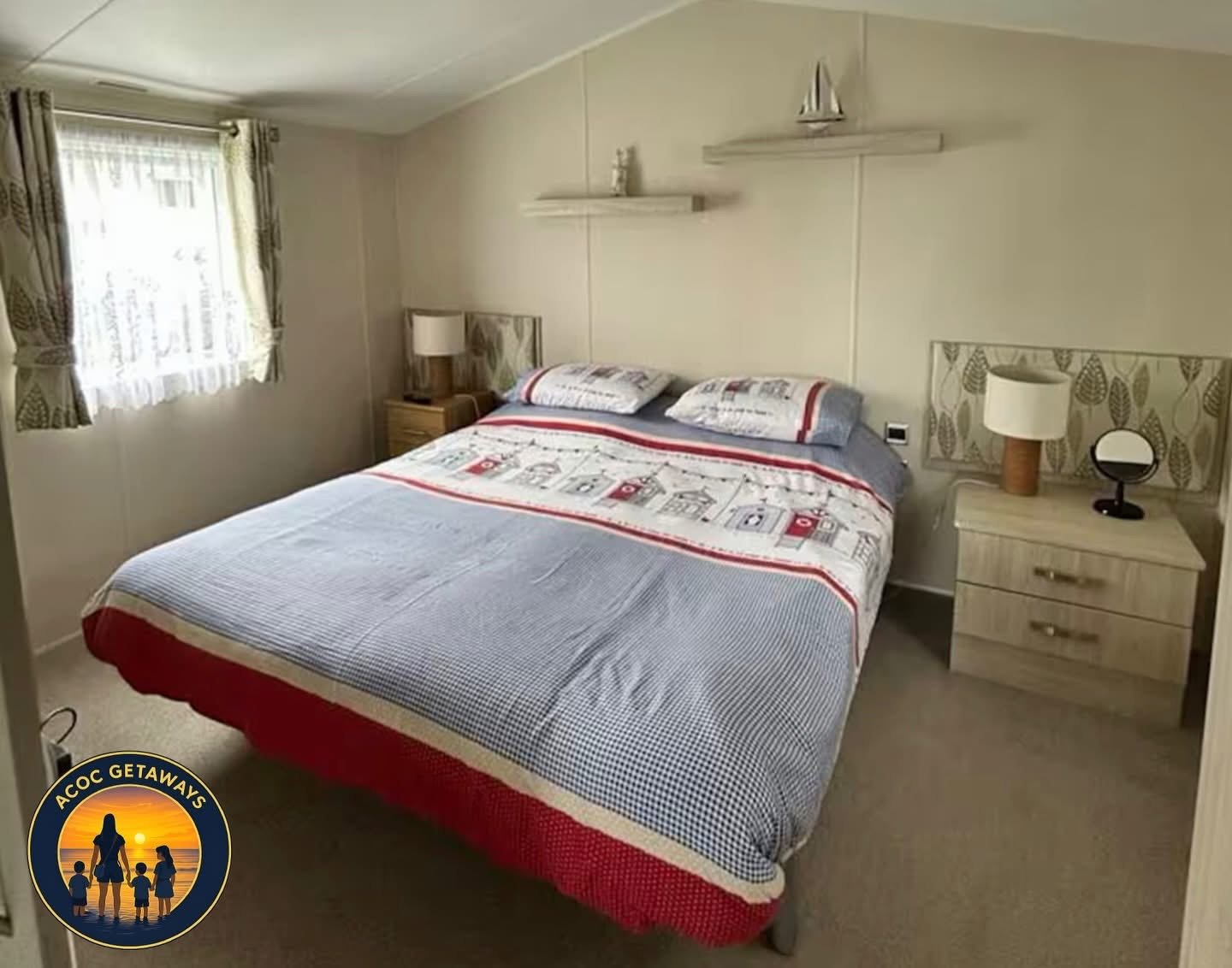 A bedroom with a bed, two nightstands with lamps, and a window with curtains. The bed has a patterned bedspread and pillows, and there is a small decorative sailboat on one nightstand.