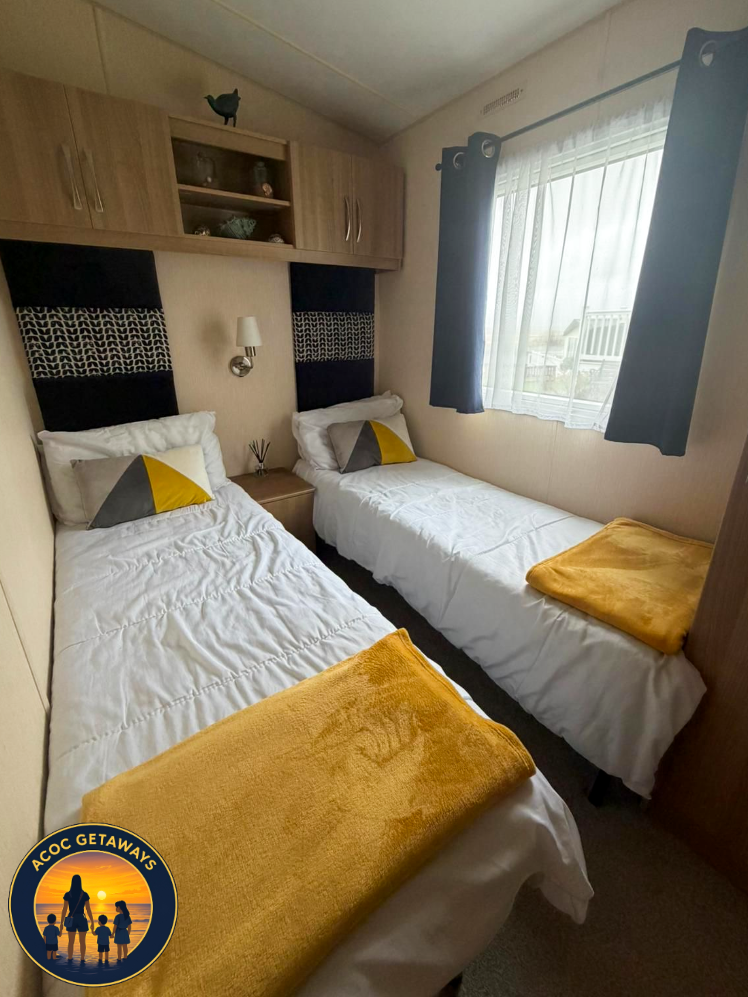 A small bedroom with two twin beds with white bedding and yellow blankets. Each bed has a triangle-patterned pillow and is against a wall with black and patterned headboards. A window with sheer white curtains and dark drapes is visible, with a woode