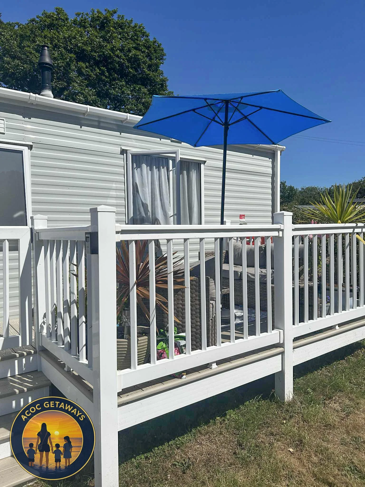 A white deck attached to a white house with a window and curtain, a blue patio umbrella, outdoor furniture, and potted plants, under a clear blue sky.