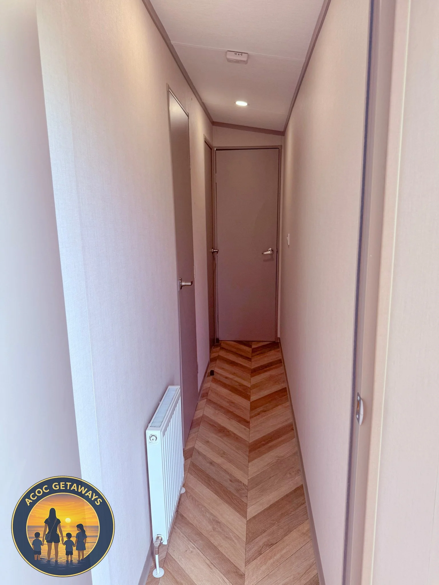 A narrow hallway with beige walls, wooden flooring, and a radiator on the left. At the end, there is a closed gray door. The ceiling has a rectangular light fixture and a smoke detector. There is an 