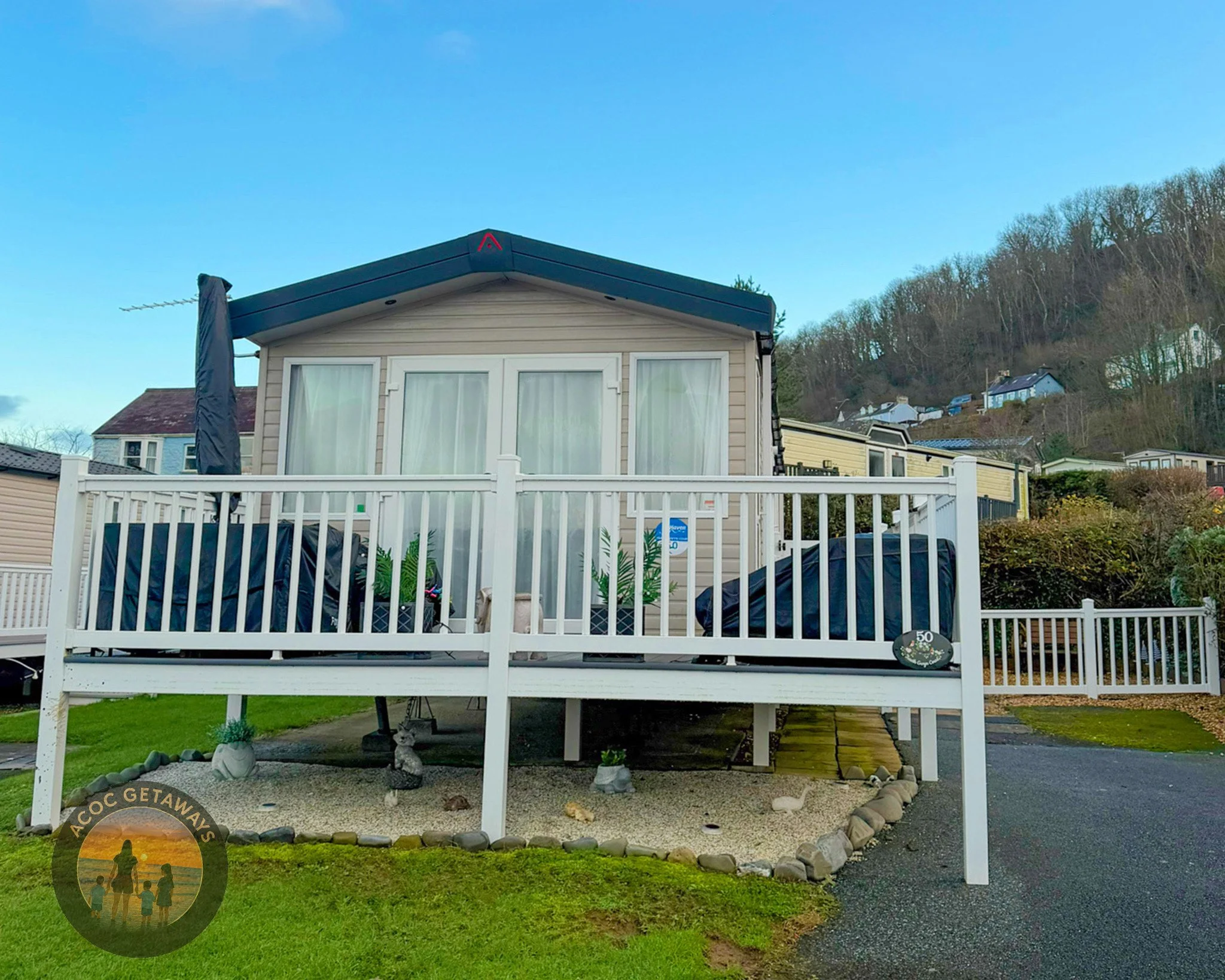 A small beige house on a raised white deck with white railing, with a view of neighboring houses on a hillside in the background, a small garden area underneath, and a clear blue sky.