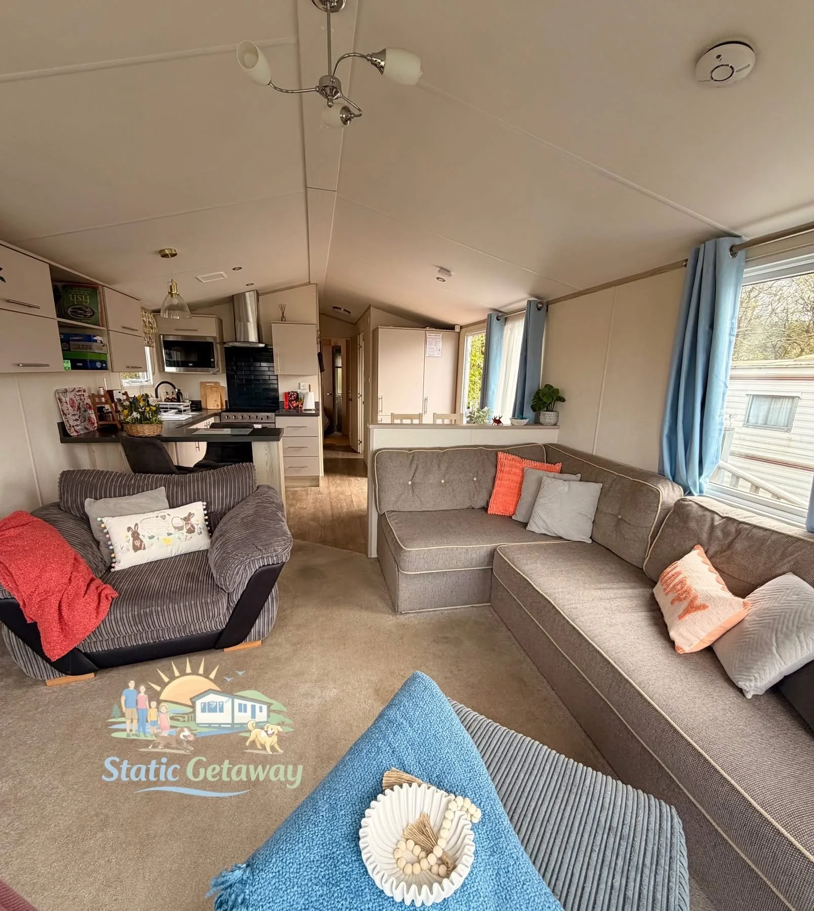 Living room with a sectional sofa, armchair, and a small table holding decorative items, with large windows and blue curtains, in a mobile home or cabin.