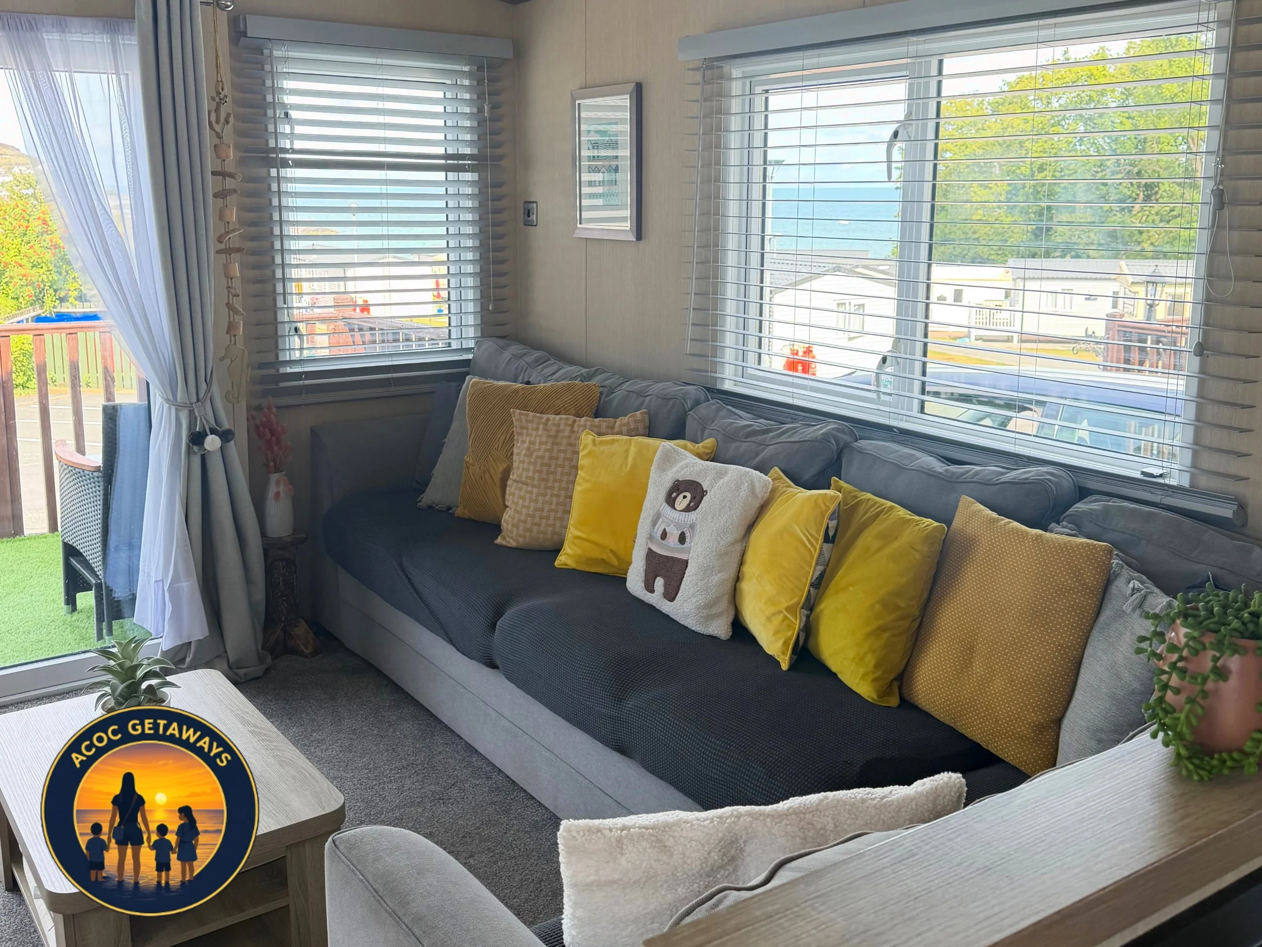 Cozy living room with large windows showing a view of the ocean and trees, featuring a gray sofa with yellow and bear-themed pillows, a glass door leading to a balcony, potted plants, and a picture on the wall.