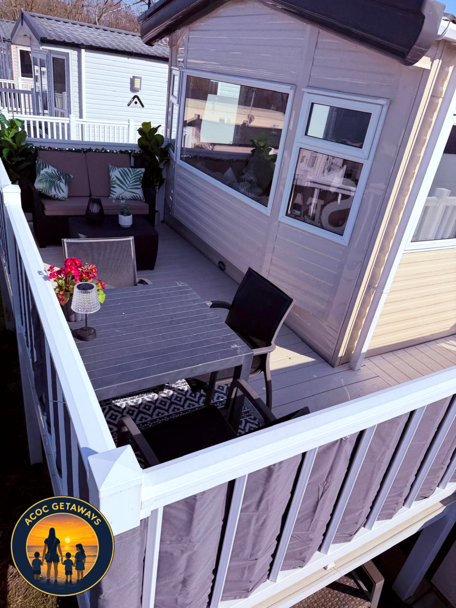 A small rooftop patio with outdoor furniture, including a loveseat with patterned pillows, a table with a flower arrangement, and a chair with a laptop. The patio is enclosed by white railing, attached to a tiny house with windows and gray siding. Th