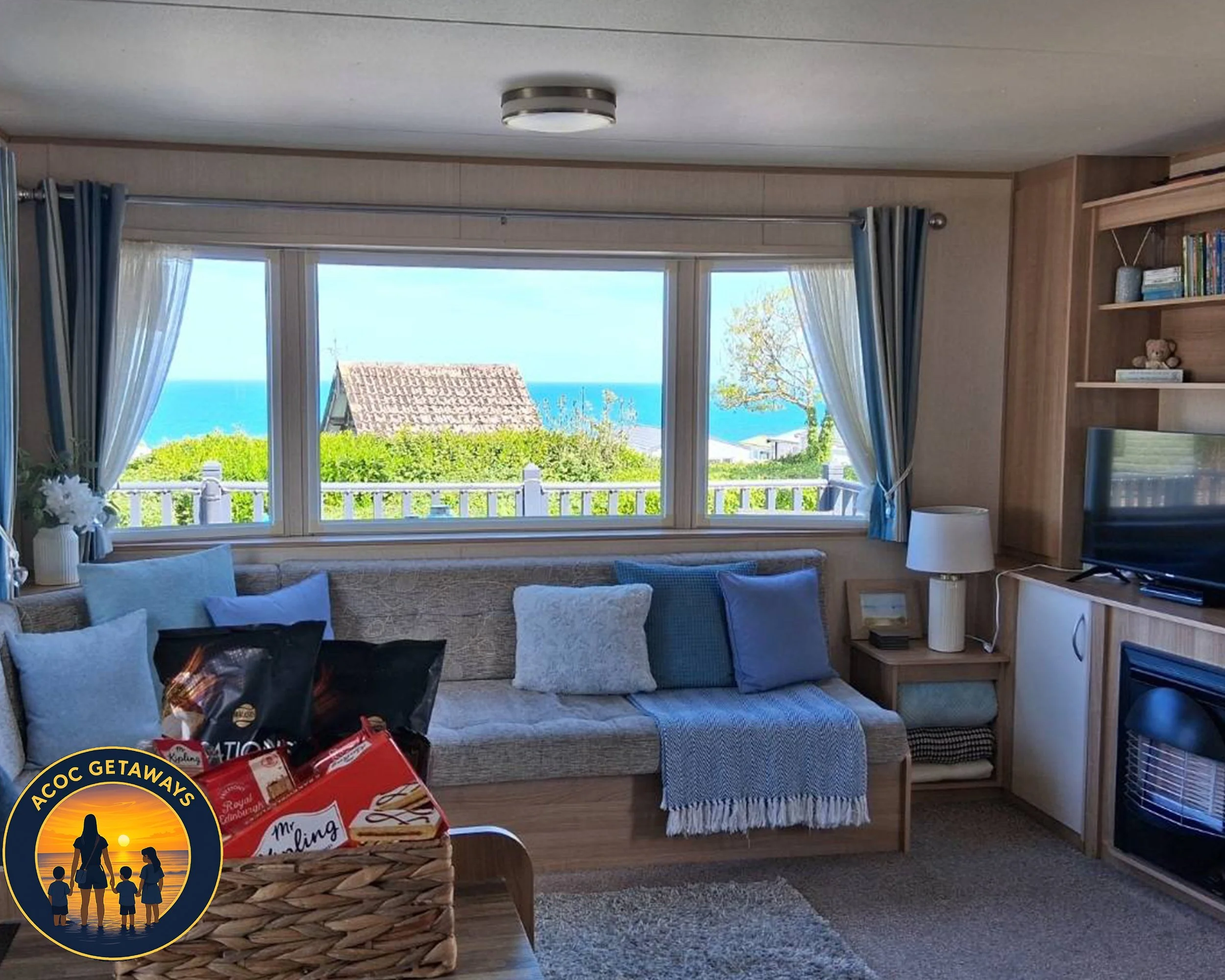 Living room with a large window view of the ocean, a beach house roof, and greenery, decorated with a sofa, cushions, a lamp, a basket with snacks, and a television, in a cozy home setting.