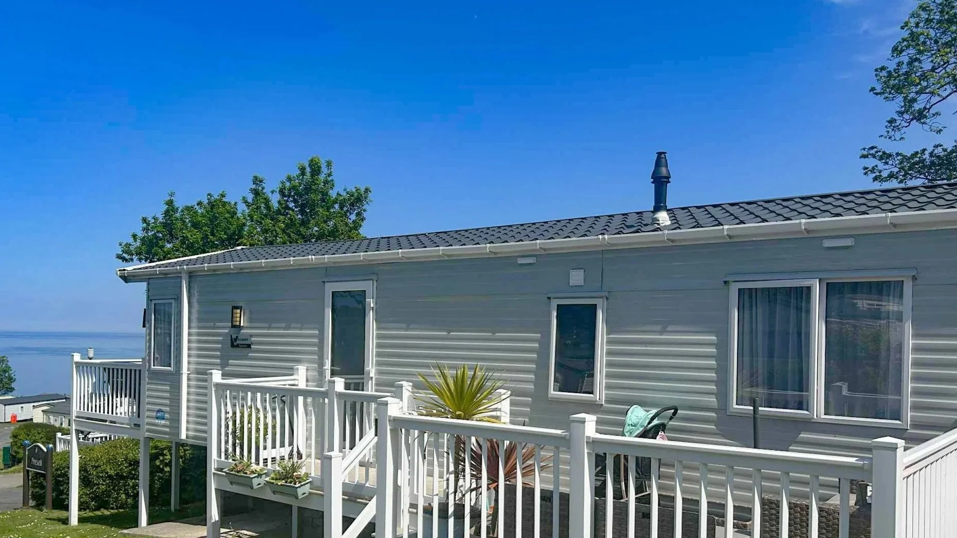 A coastal house with a grey exterior, white railing balcony, palm plant, and a view of the ocean in the background under a clear blue sky.