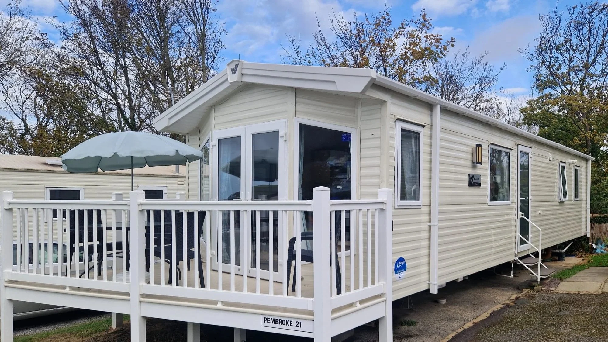 White mobile home with a deck and railing, outdoor table with chairs, umbrella, surrounded by trees with fall foliage, and a partly cloudy sky.